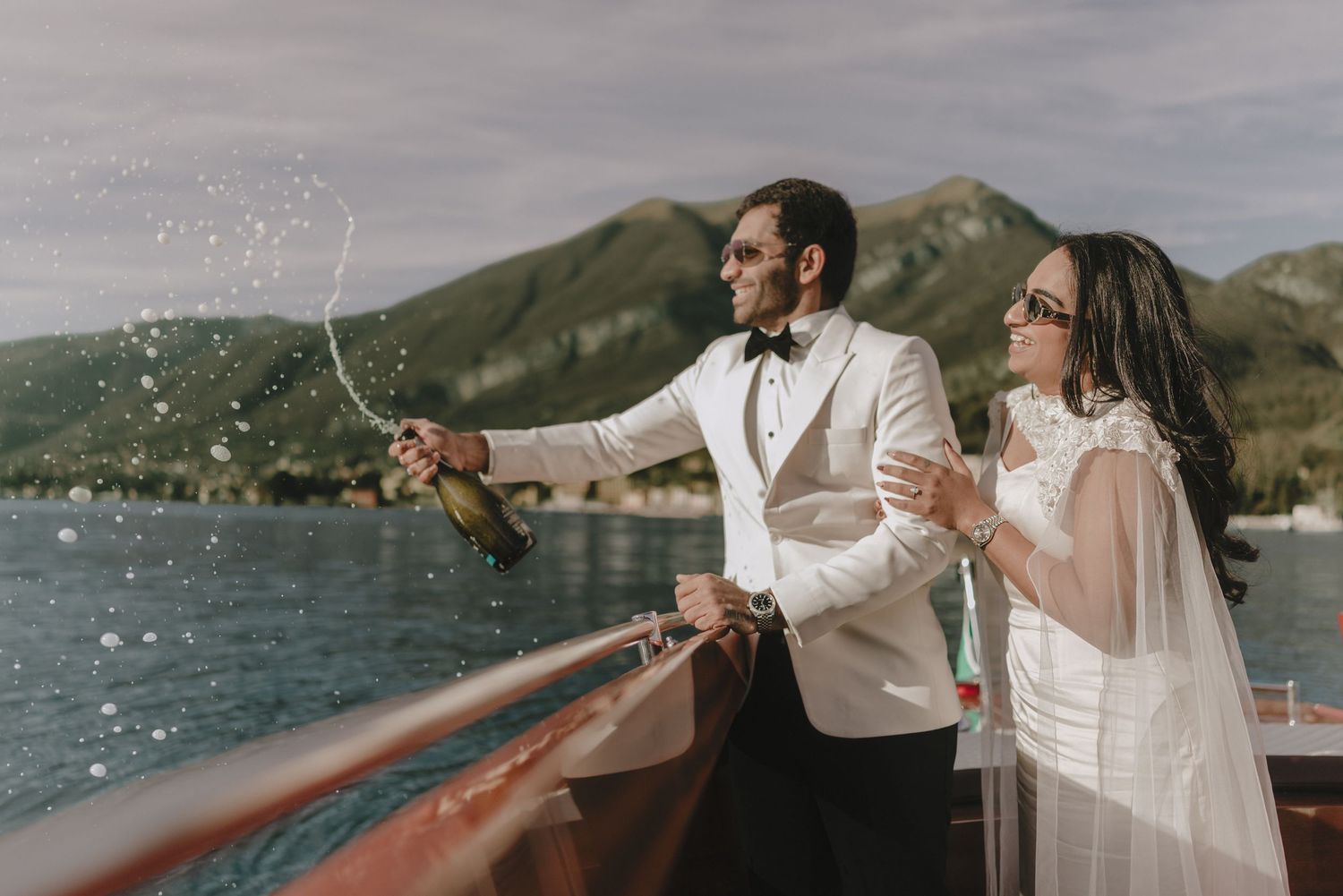 Bride and groom celebrating on a boat, popping champagne with mountain lake backdrop on their wedding day.