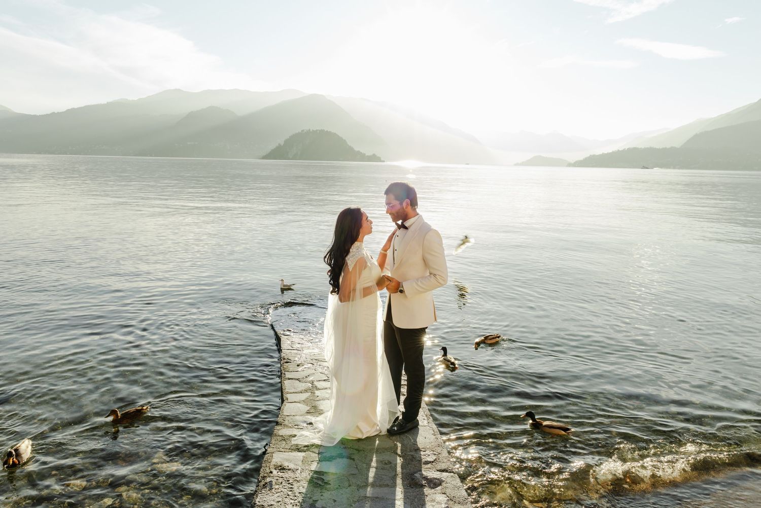 Bride and groom standing on a wooden dock at golden hour with serene mountain lake backdrop and ducks nearby.