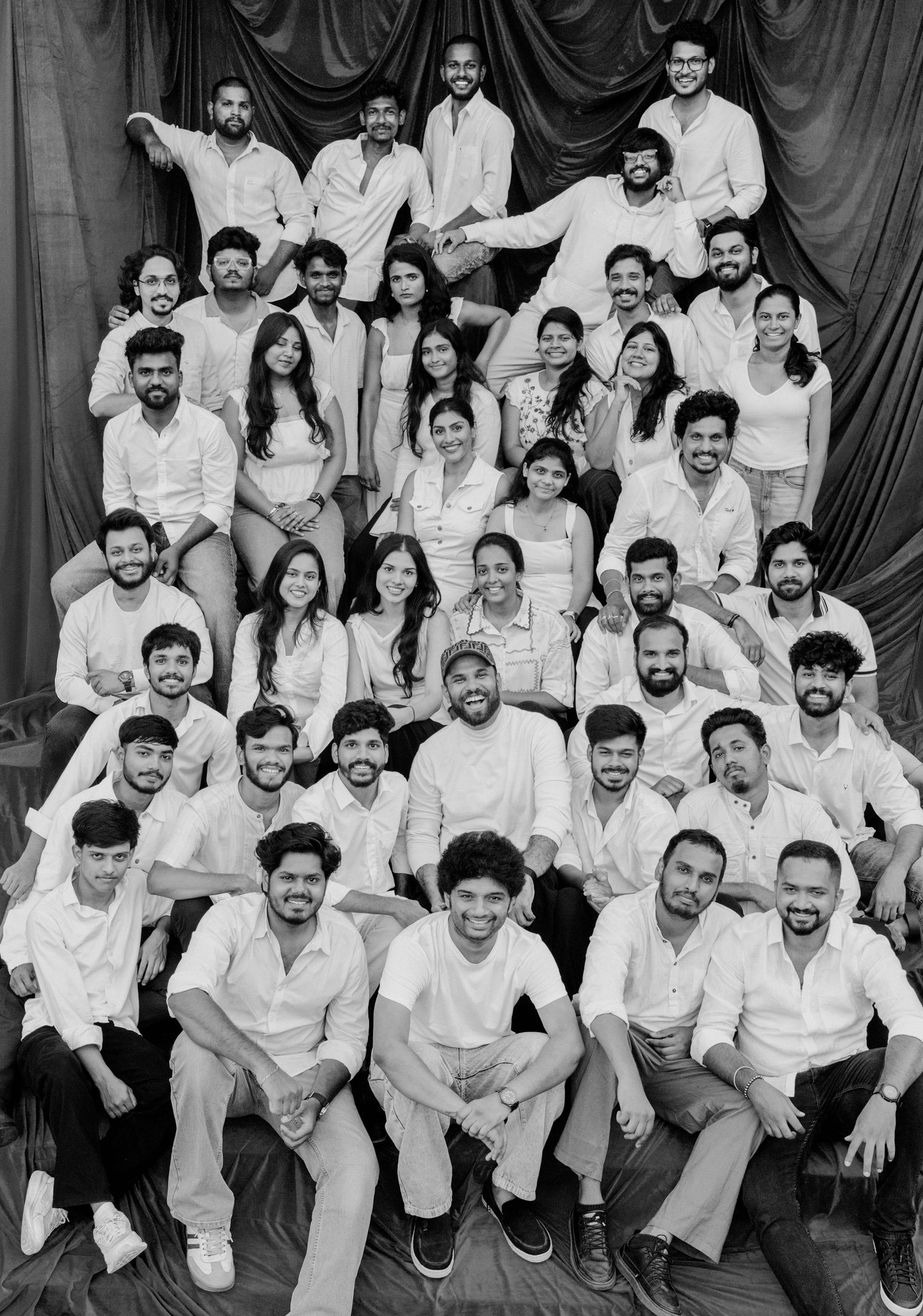 Black and white group photo of diverse young adults in white outfits posed in pyramid formation against draped backdrop.