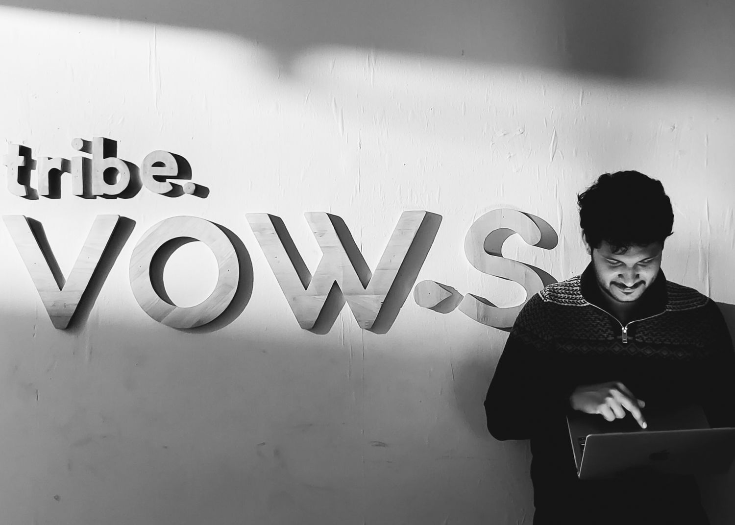 Black and white photo of a man smiling at laptop in front of tribe.VOWS wall sign.