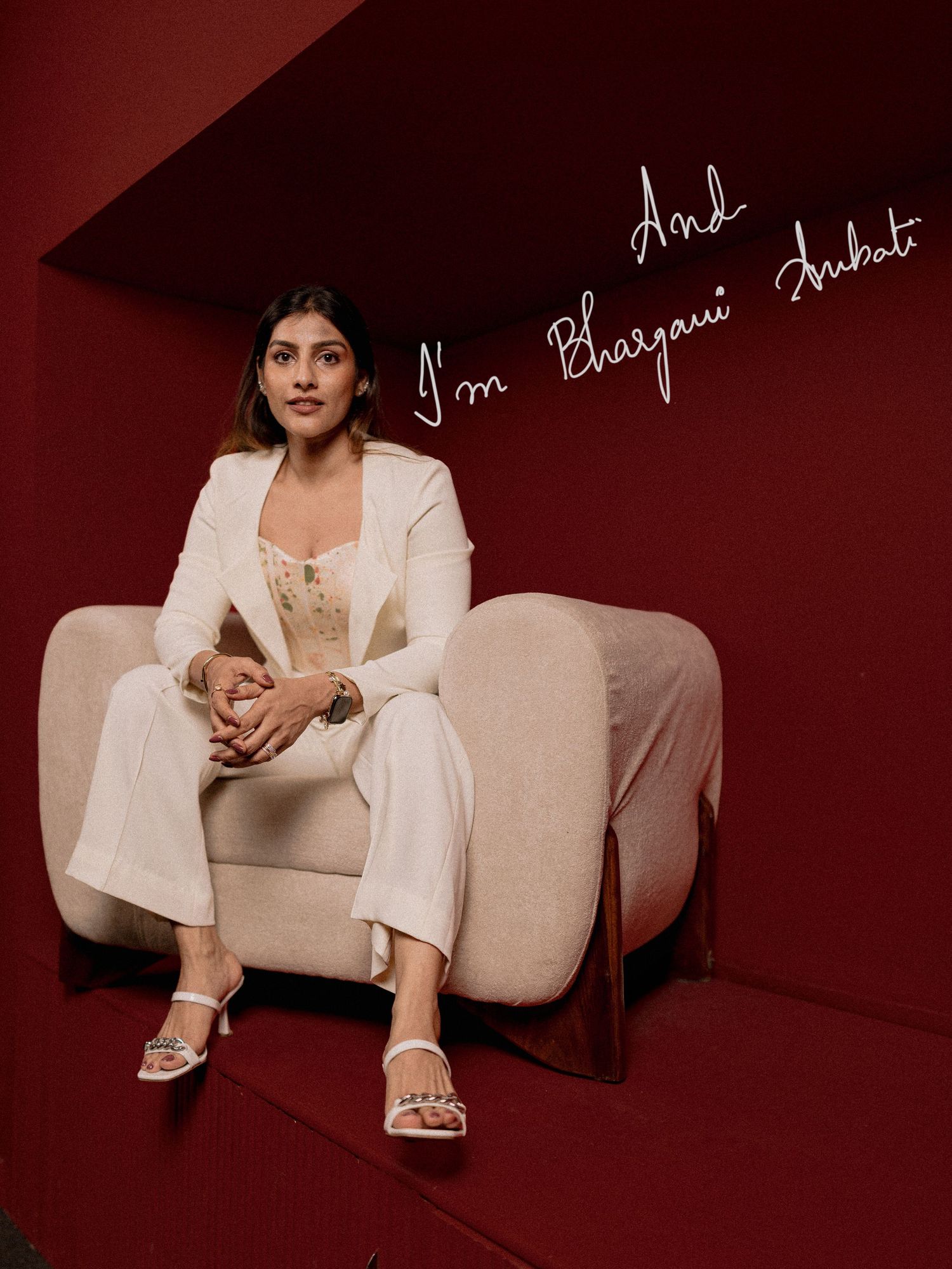 Bhavyami Ambati sits elegantly in a cream blazer and wide-leg trousers on a modern chair against a red wall.