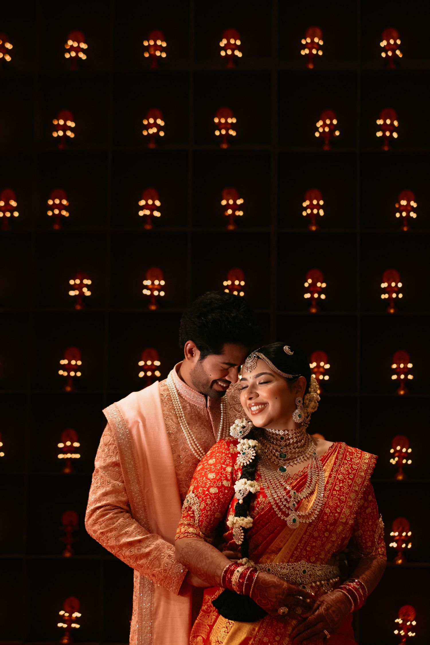 Indian wedding couple in coral and orange traditional attire embrace in front of a dark wall glowing with rows of warm red lanterns.