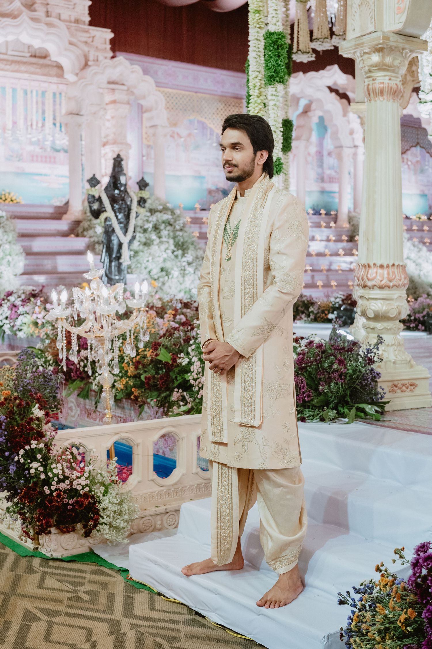 Groom in cream embroidered sherwani stands barefoot inside elaborately decorated mandap with floral arrangements.
