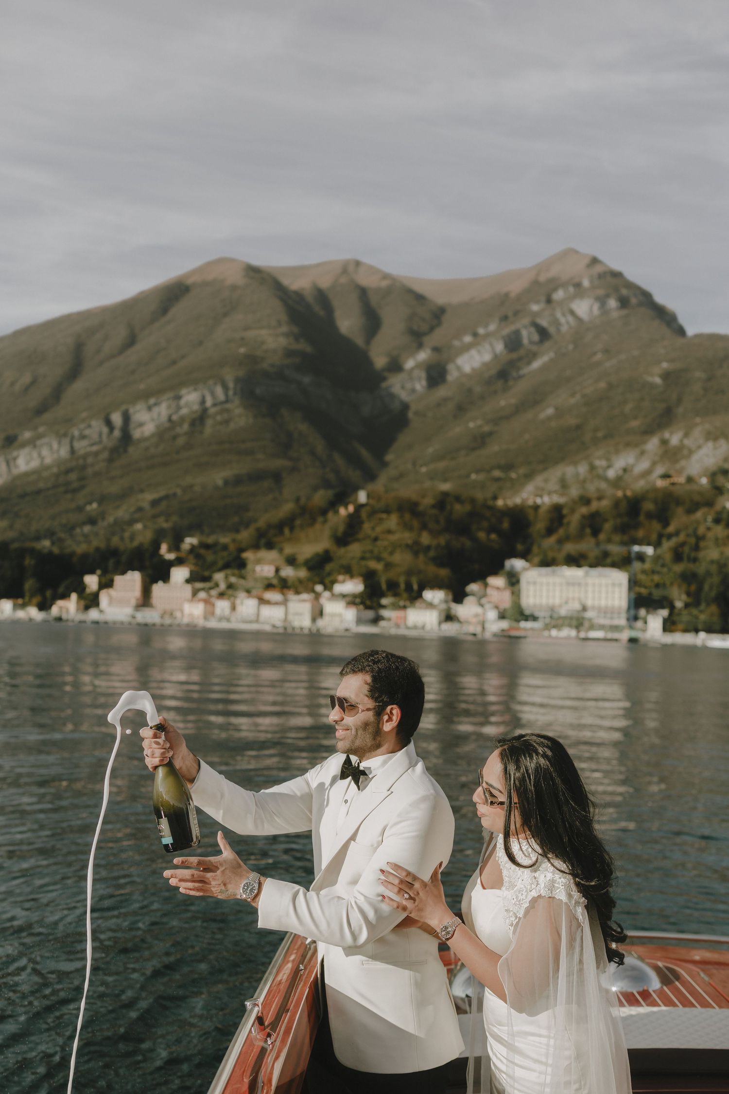 Newlyweds celebrate with champagne on a wooden boat with stunning Lake Como mountain backdrop.