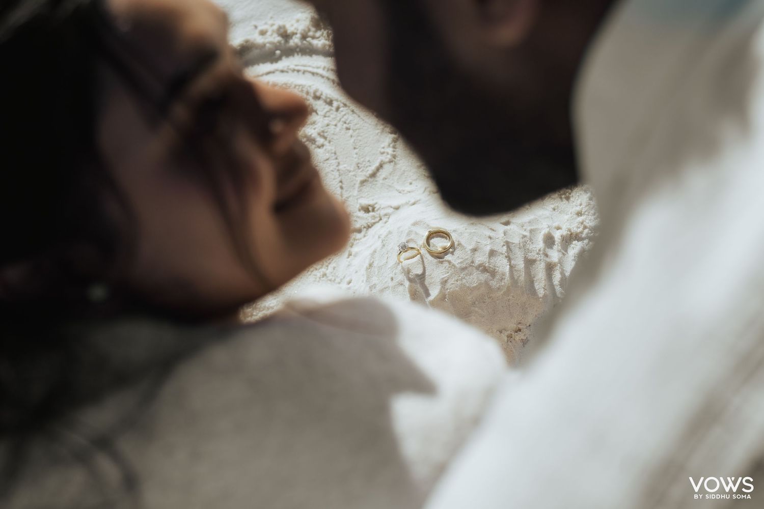 Intimate wedding couple lying together with gold rings in soft focus foreground, romantic bridal photography.