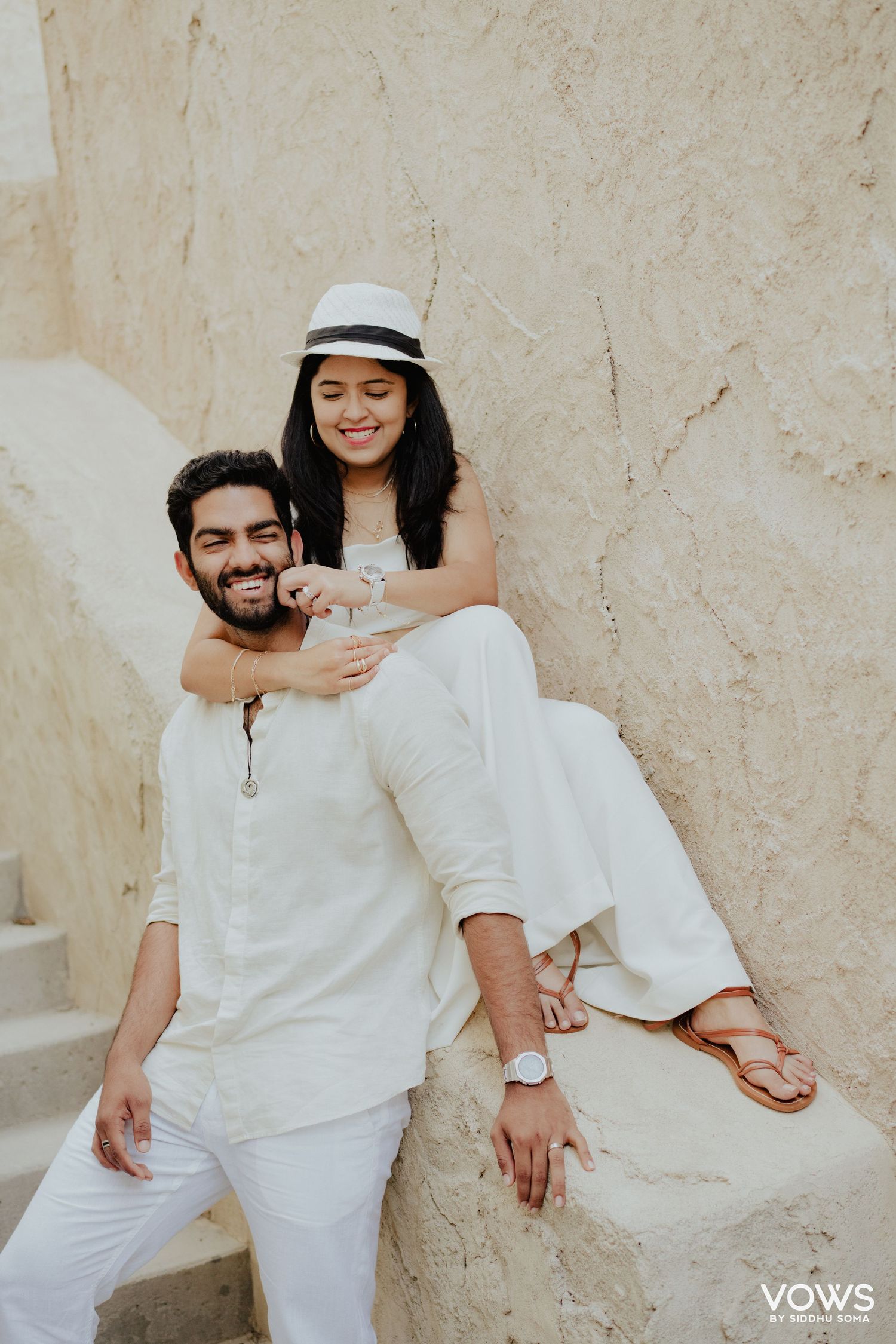 Couple in matching white outfits posing on stone steps, woman in hat sitting above smiling man.