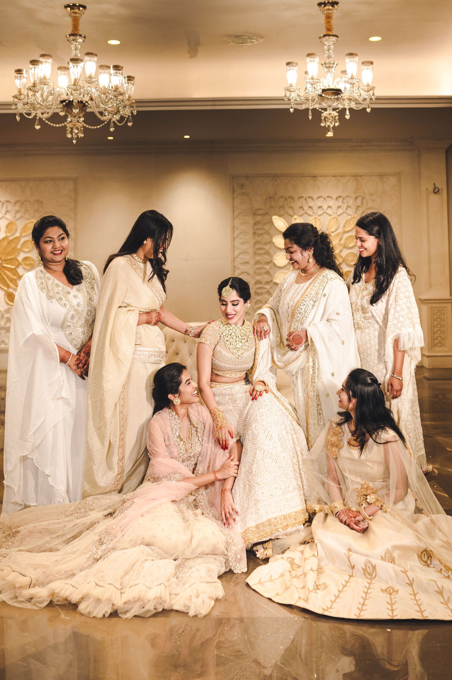Bride in white lehenga surrounded by bridesmaids in ivory outfits under chandeliers at an elegant Indian wedding.