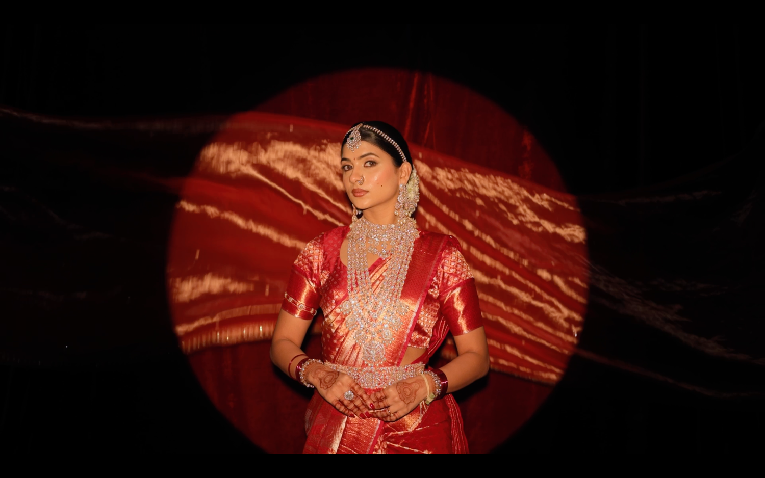 A bride in a red silk saree and traditional jewelry poses elegantly against a dramatic circular red backdrop.