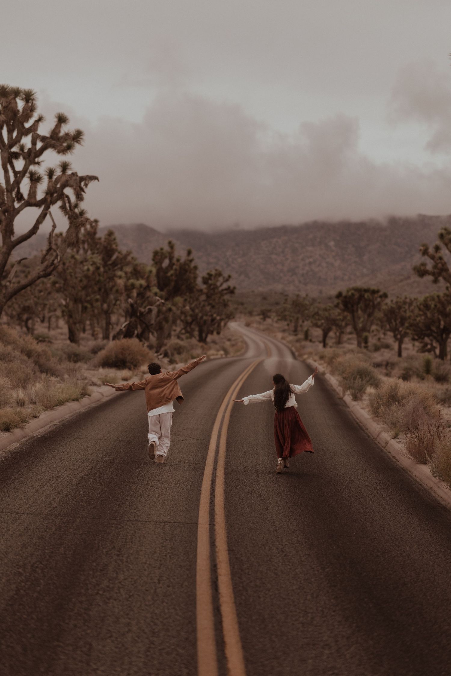 Two people walk hand-in-hand down a desert road lined with Joshua trees under a moody, overcast sky.