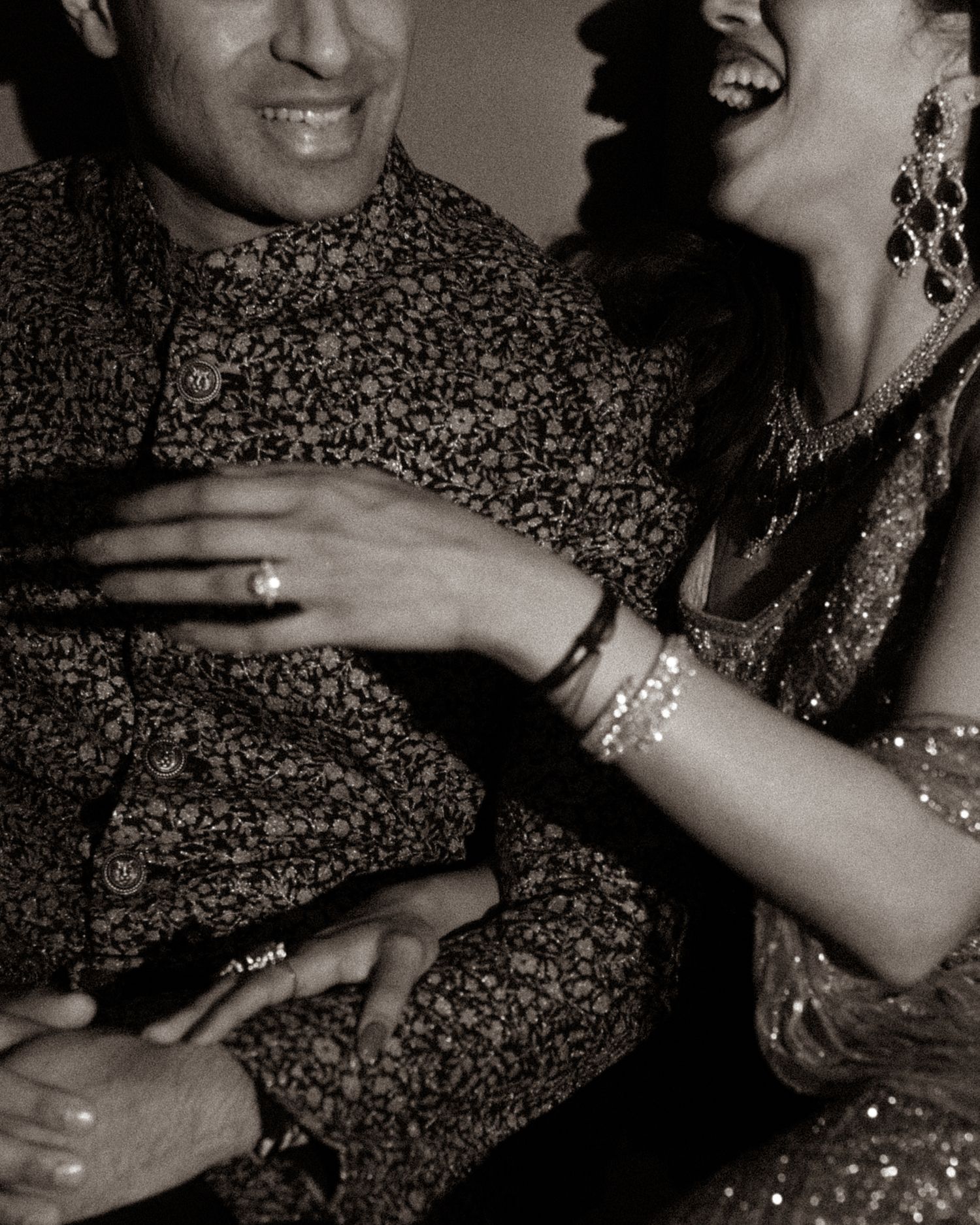Laughing couple in traditional Indian attire, woman wearing gold jewelry and bangles, man in floral kurta, black and white photo.