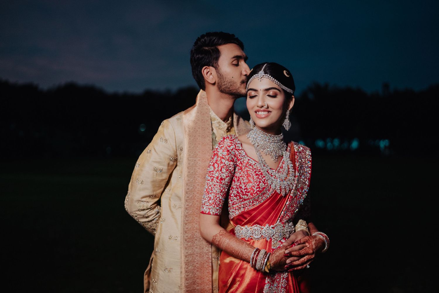 Indian couple in traditional wedding attire, bride in red saree with jewelry, groom in gold sherwani, posing outdoors at dusk.