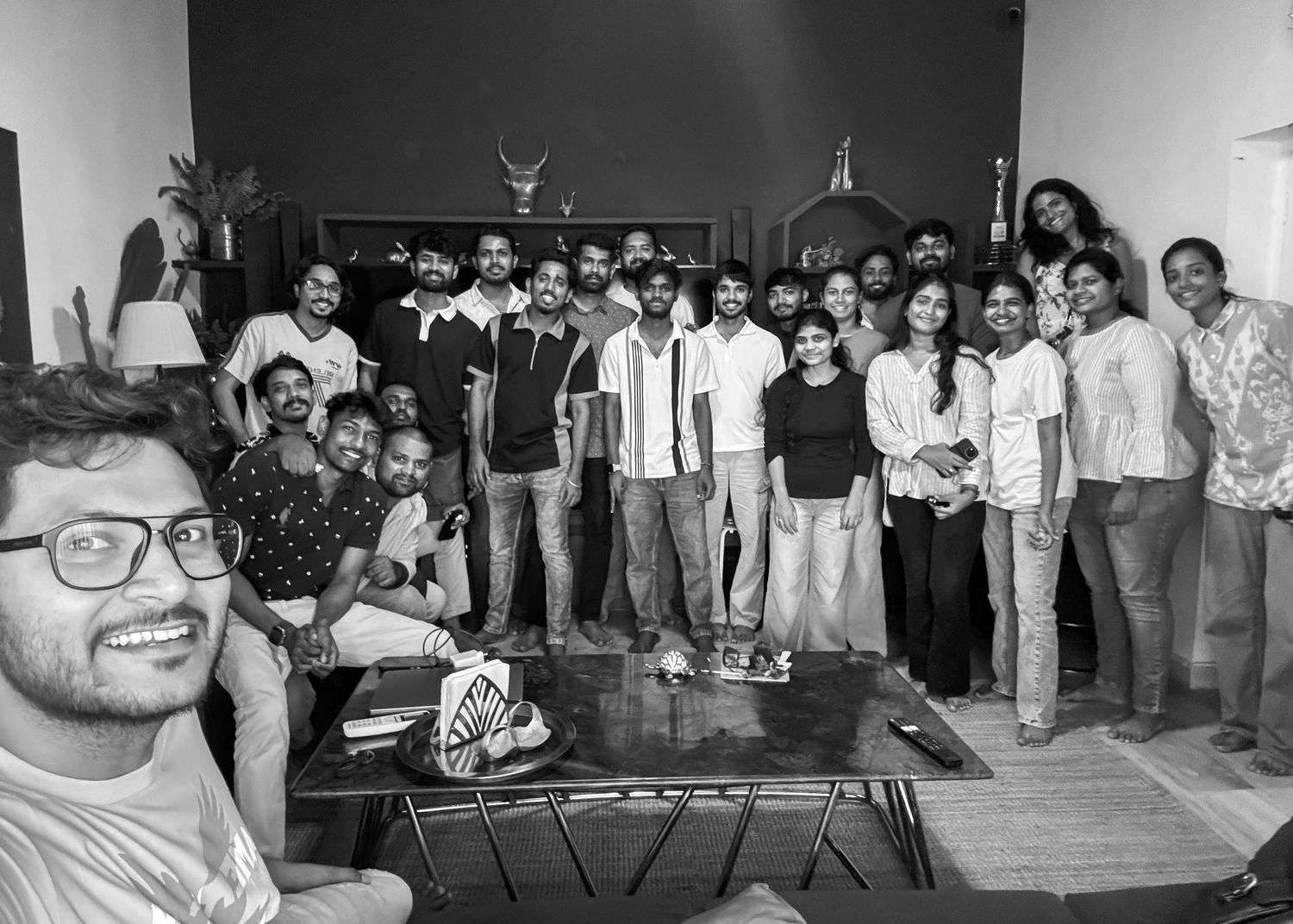 Black and white group photo of young adults posing together indoors for a selfie in a casual setting.