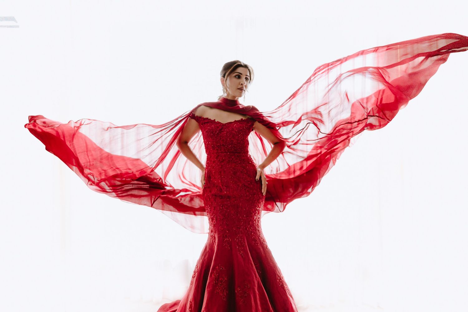 Woman in stunning red mermaid gown with flowing fabric wings against white background.