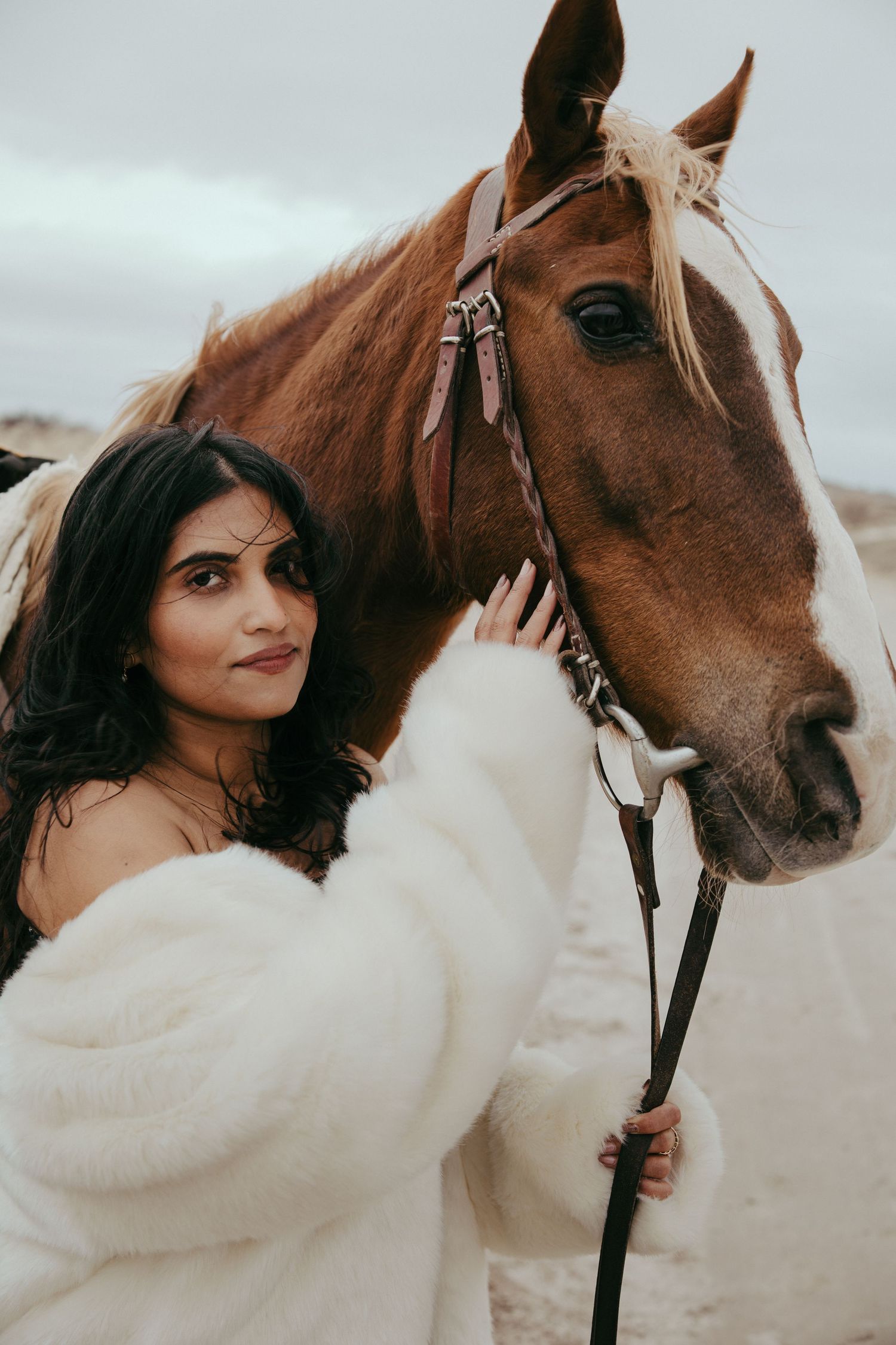 A woman in a white fluffy coat stands beside a brown and white horse, gently holding its reins outdoors.