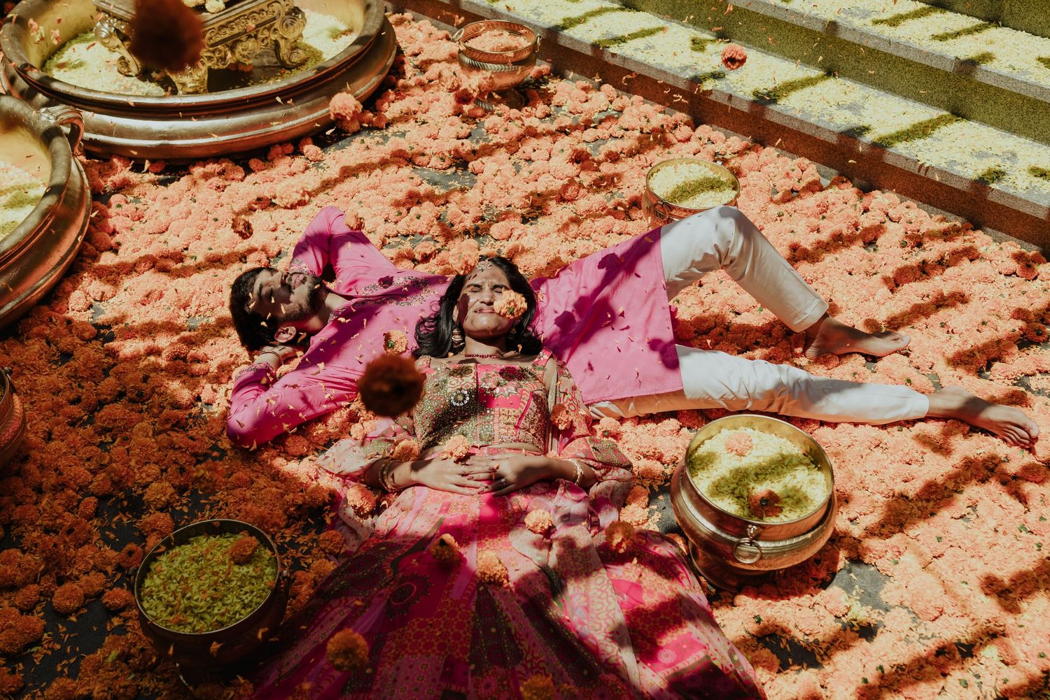 Indian bride and groom lying on a floral carpet surrounded by traditional wedding ceremony items.