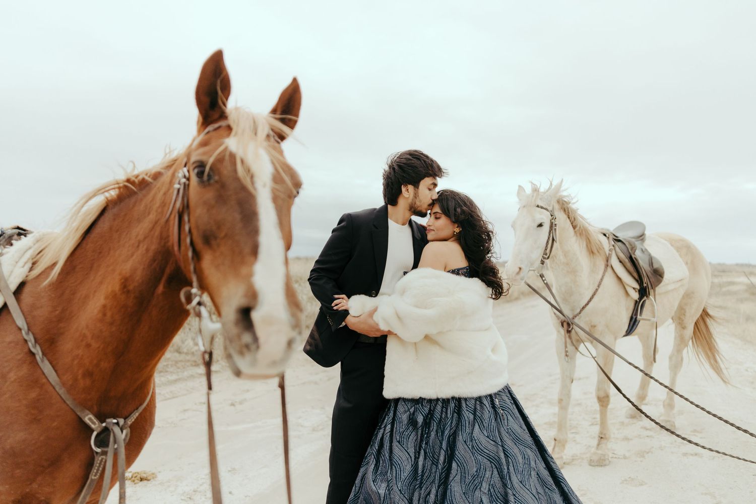 Romantic couple in formal attire sharing a tender moment surrounded by brown and white horses on a sandy landscape.