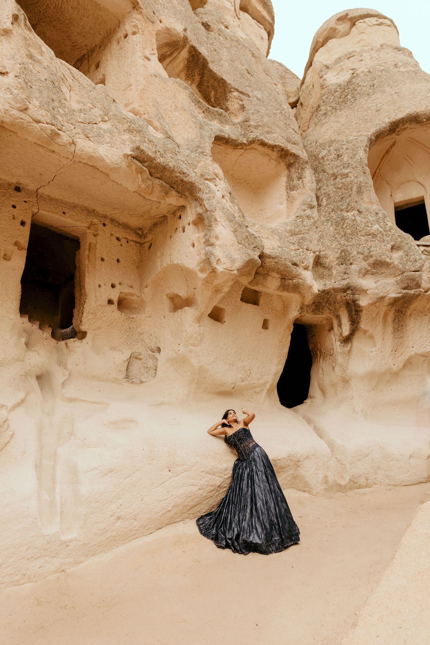Woman in black gown poses dramatically before ancient cave dwellings carved into golden sandstone rock formations.