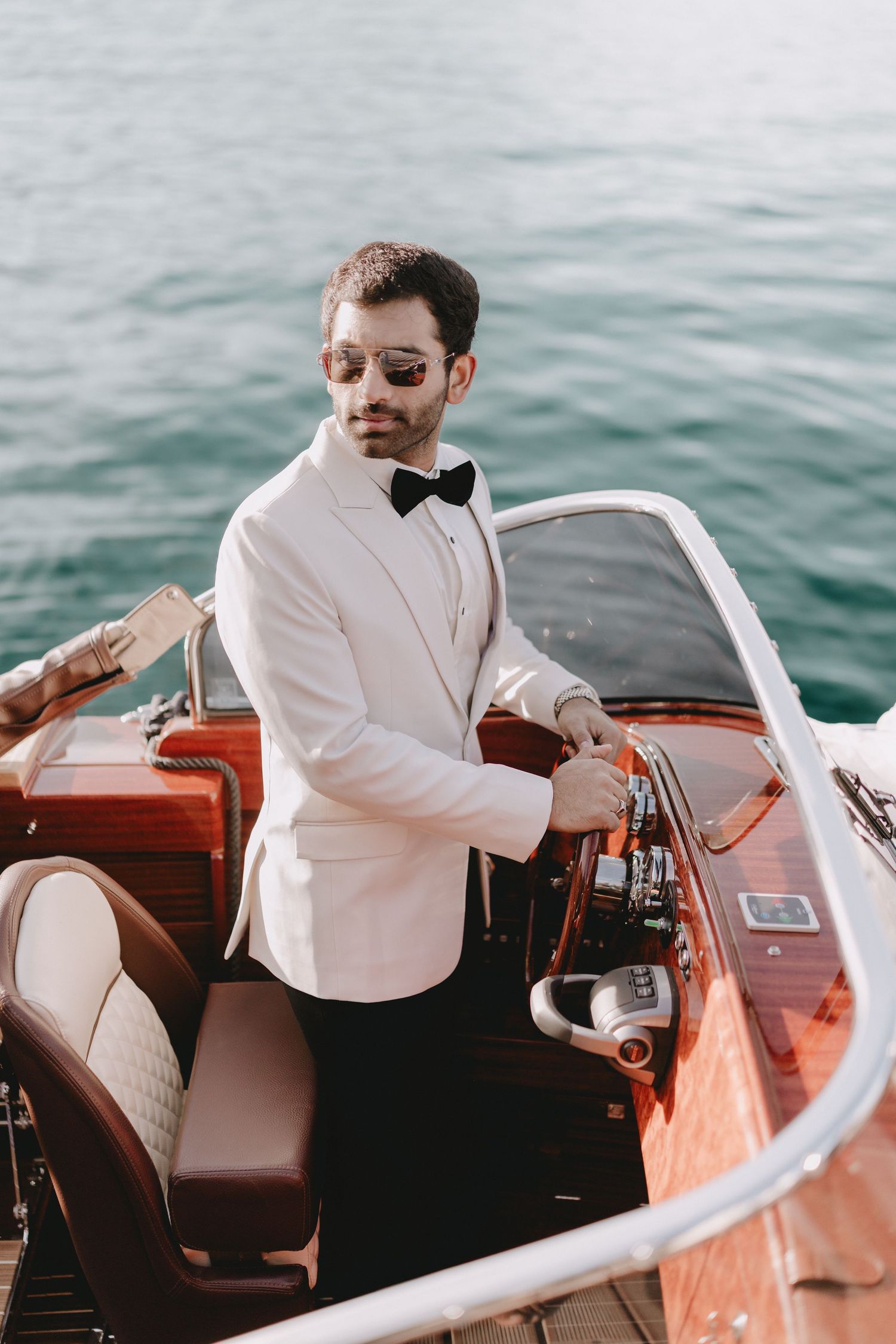 An elegant groom in a white tuxedo jacket steers a classic wooden speedboat on the water.