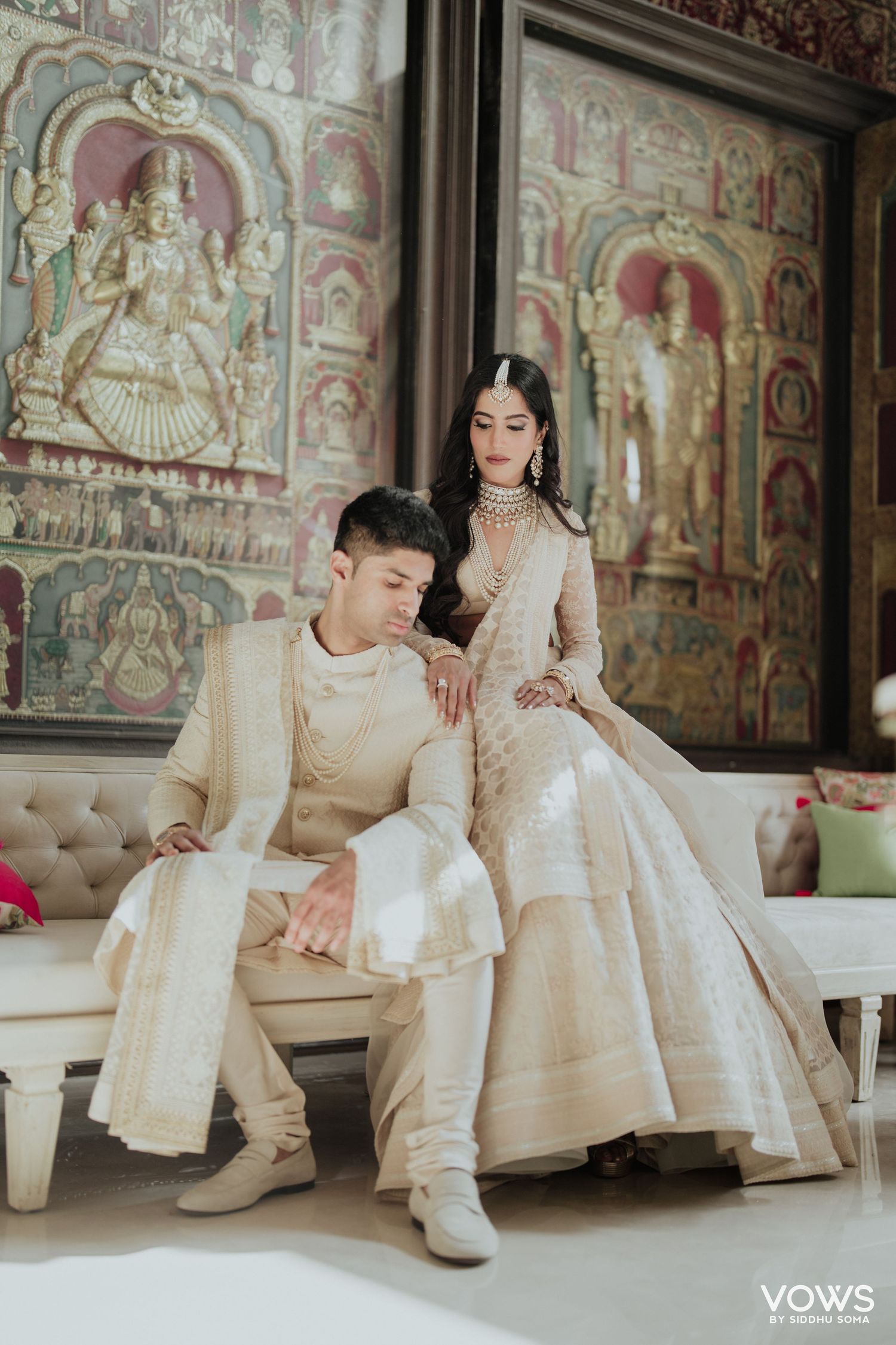 Couple in coordinating ivory Indian wedding attire posed elegantly before ornate painted temple murals.