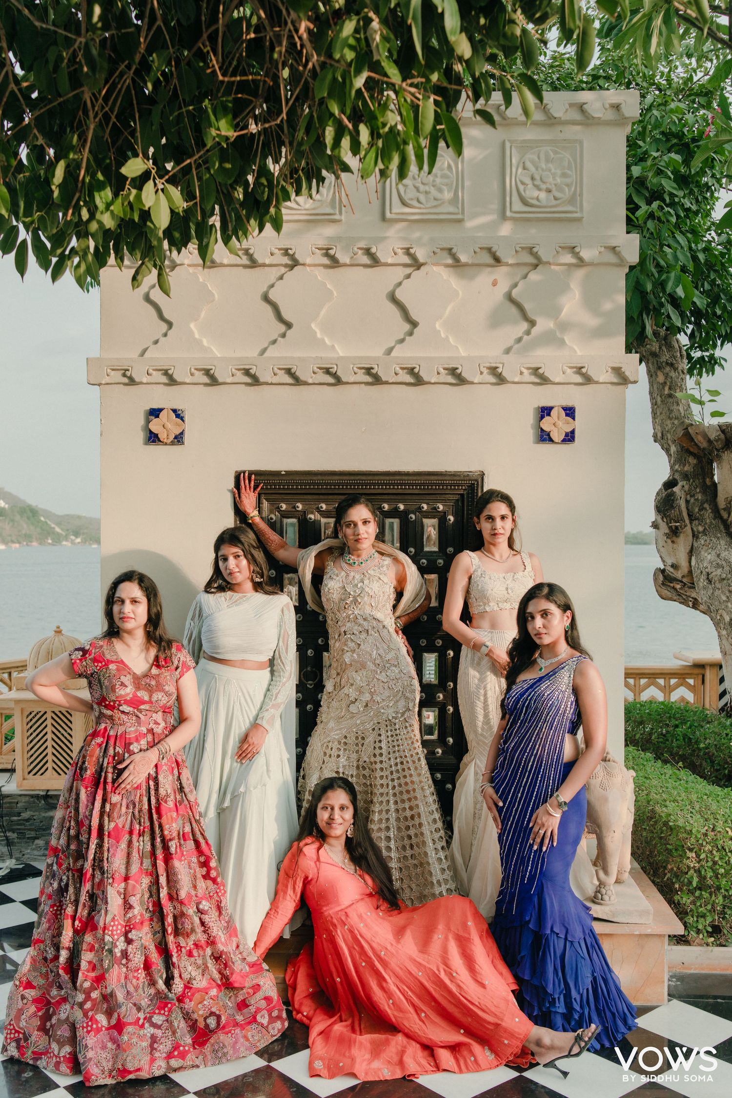 Six women in vibrant Indian ethnic wear pose elegantly at a lakeside palace venue during a wedding celebration.