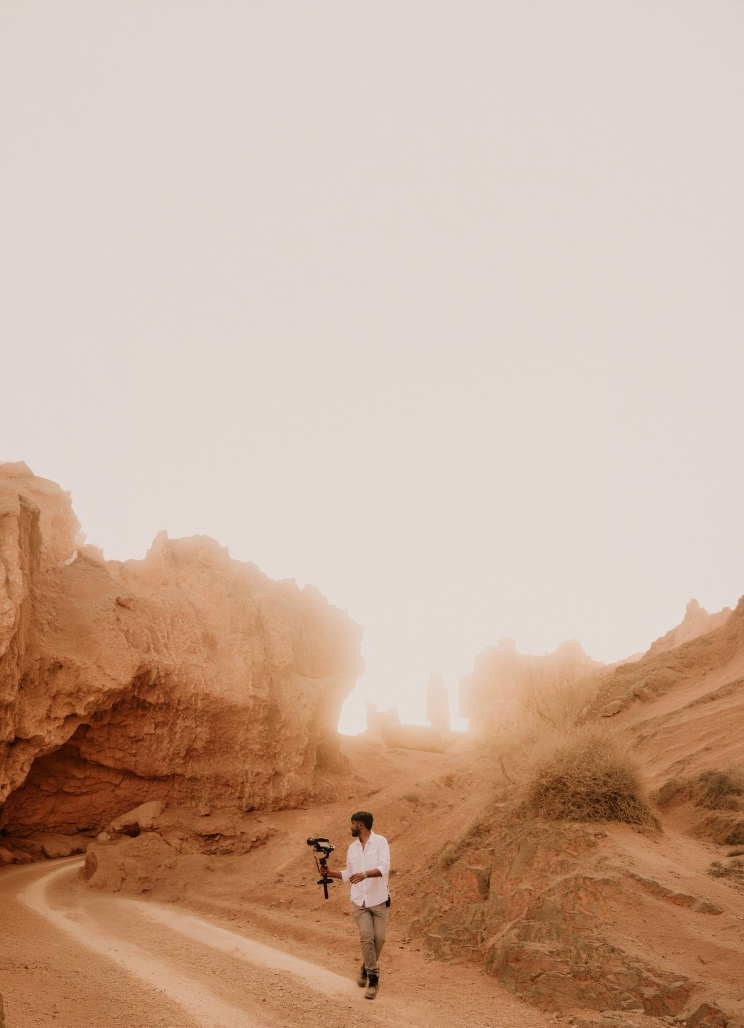 A filmmaker walks through a misty red rock canyon on a dusty trail, carrying a camera stabilizer.