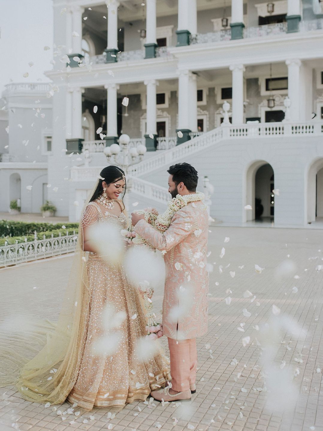 Couple in traditional Indian wedding attire stand before a grand white palace amid falling flower petals.
