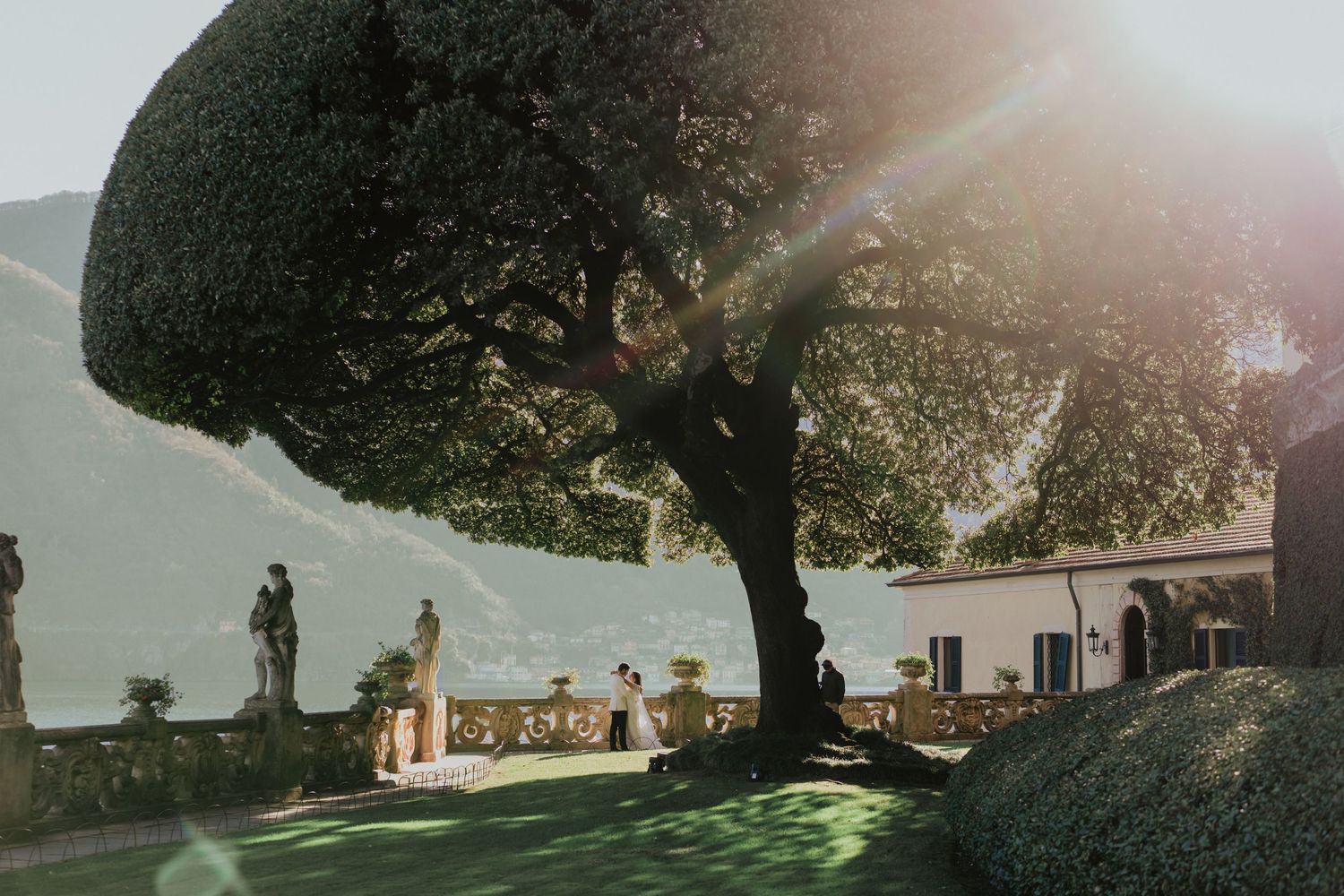 Sunlit garden terrace with a large tree, classical statues, and mountain views at an Italian villa wedding venue.