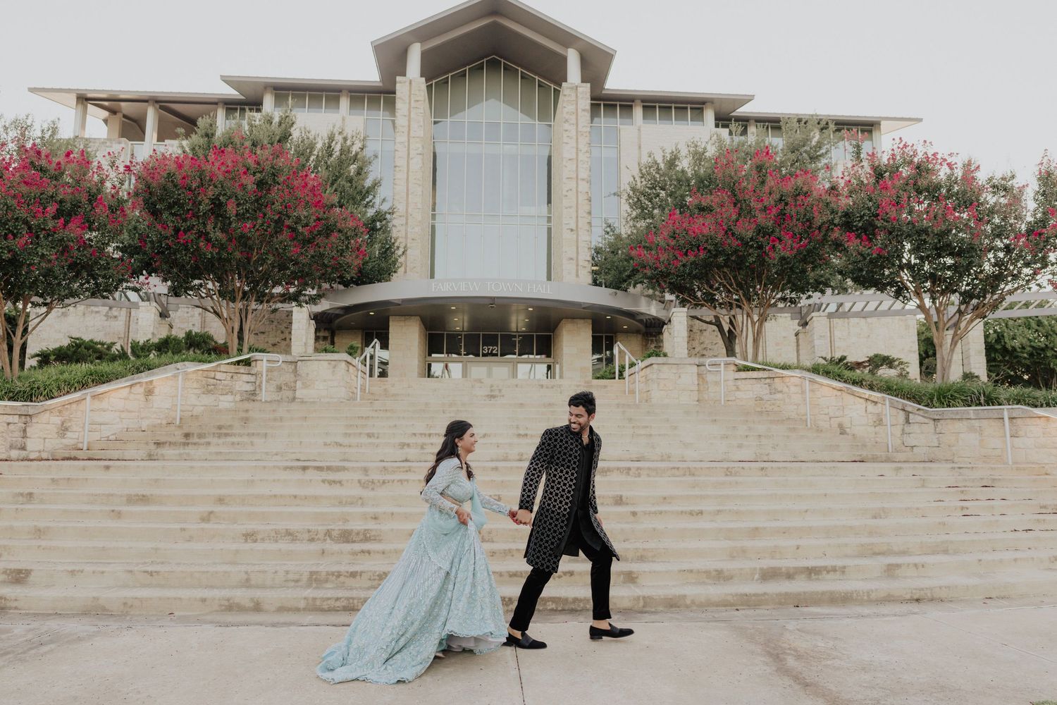 Couple in traditional South Asian attire holding hands on steps outside elegant building with pink flowering trees.
