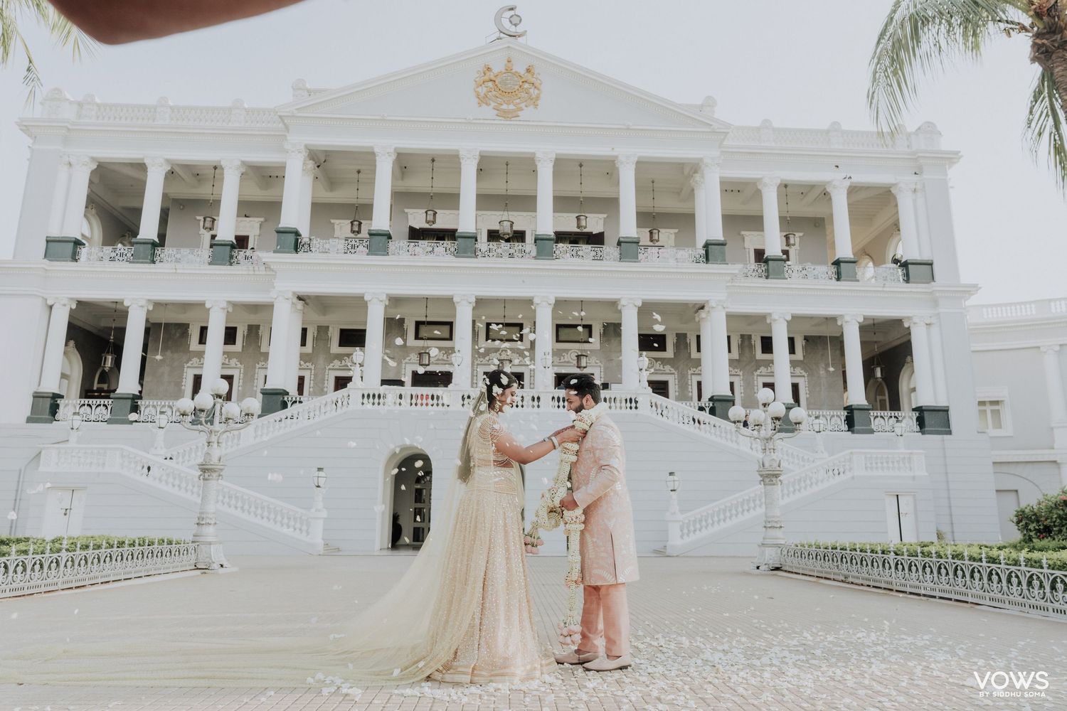 Bride and groom stand before a grand white colonial wedding venue with elegant staircases and ornate architecture.