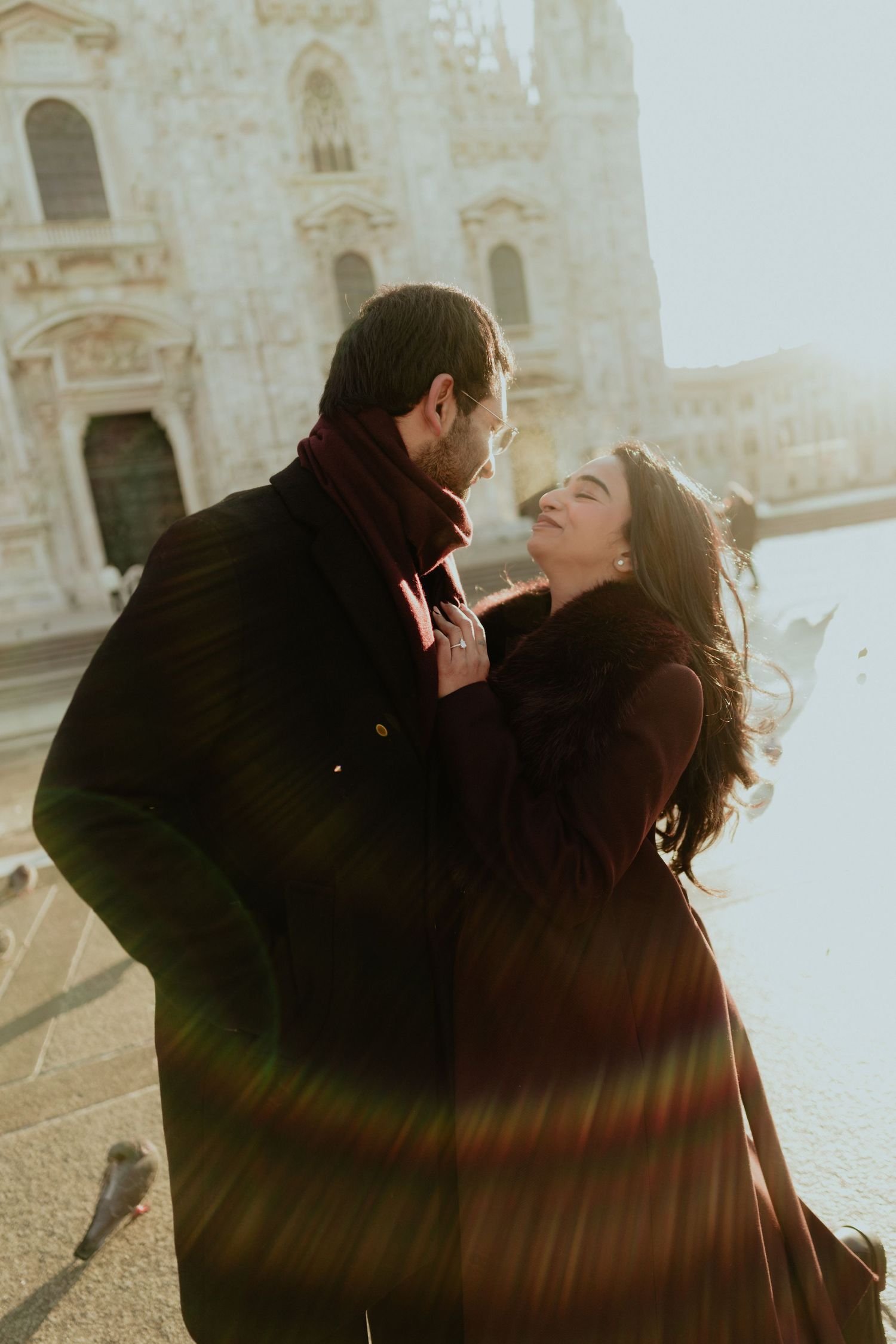 Romantic couple embraces in warm sunlight before Milan's stunning Duomo cathedral on a windy day.