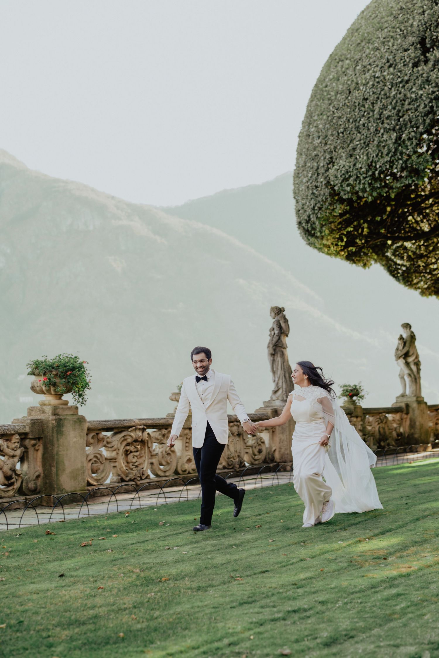 Bride and groom running hand in hand on lush lawn at elegant Italian villa with mountain backdrop.