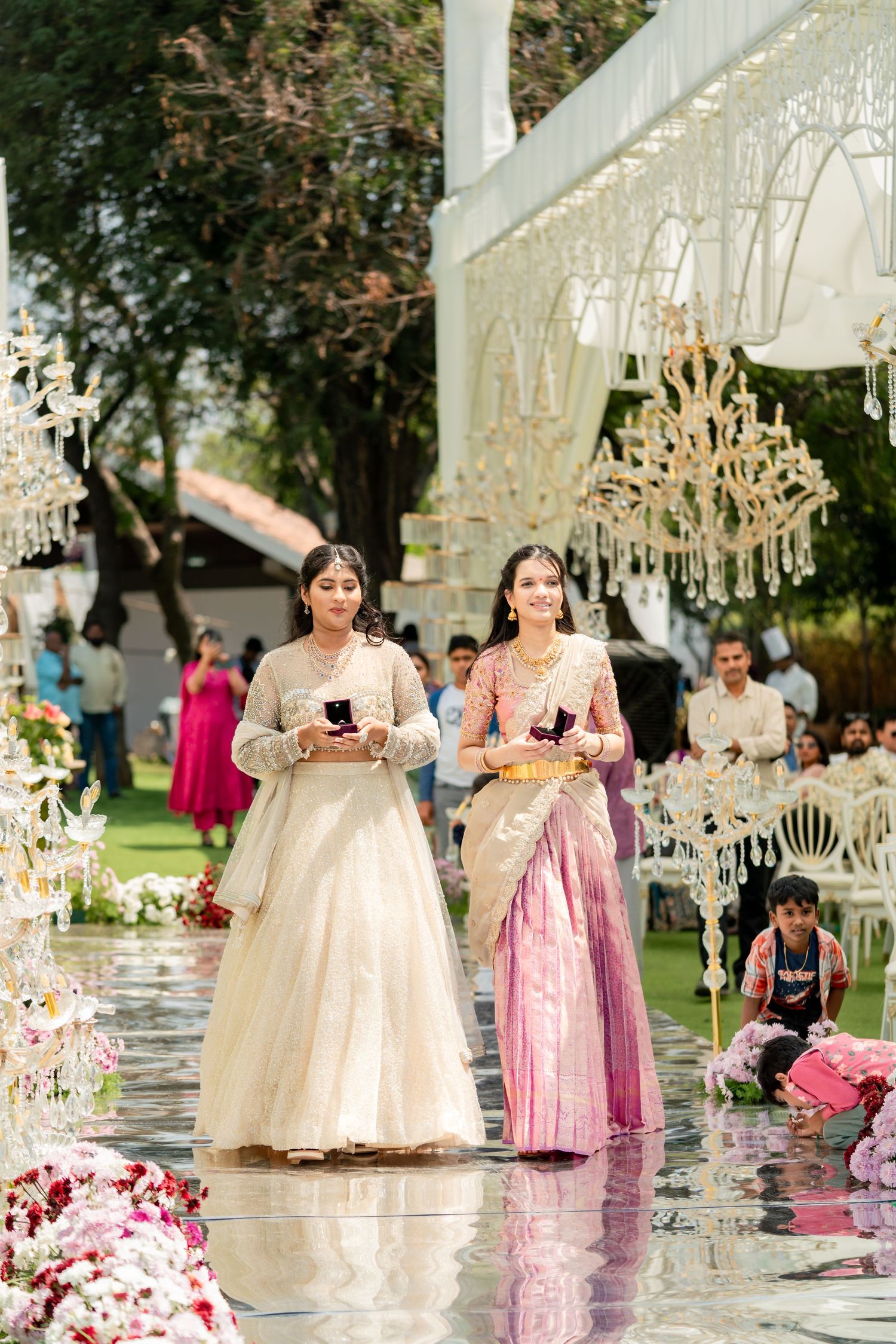 Two women in traditional Indian attire walk down reflective aisle at decorated outdoor wedding ceremony.