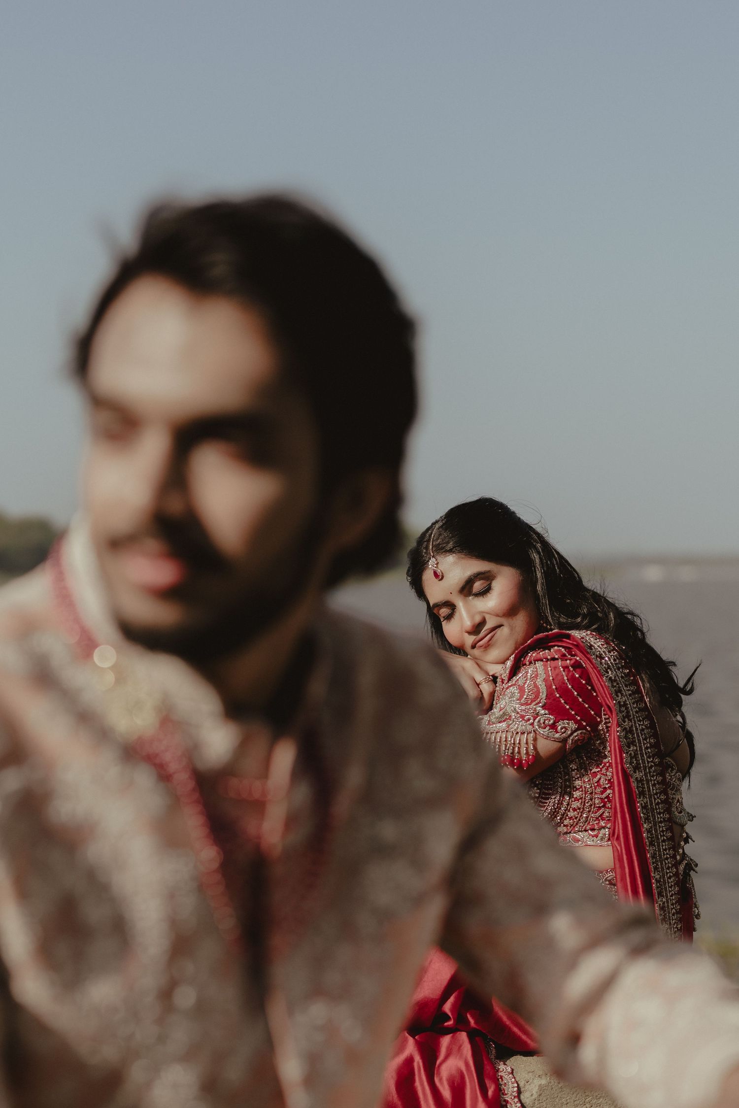 Indian bride in red lehenga leans on groom in embroidered sherwani during traditional wedding portrait outdoors.