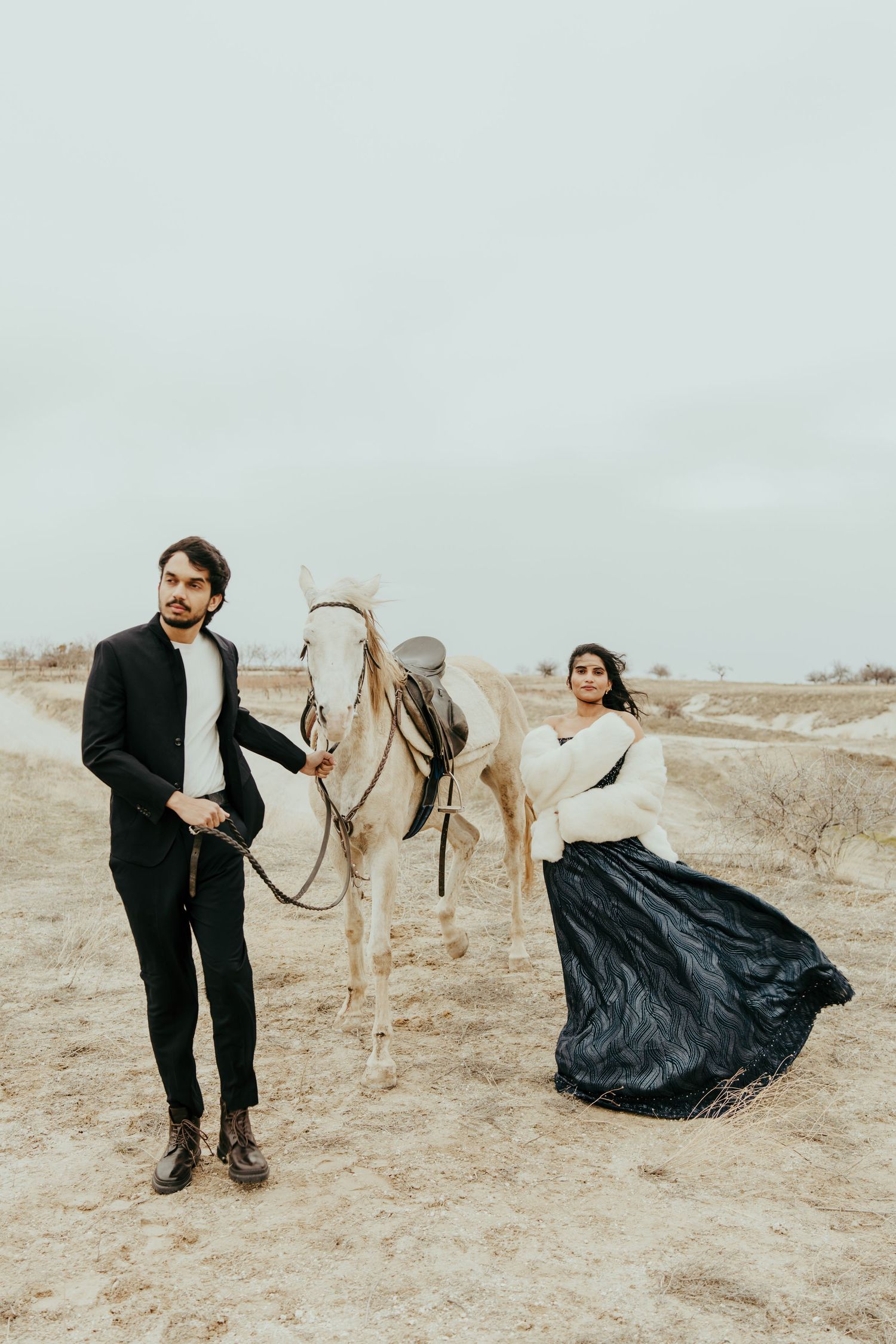 Couple in elegant attire poses with a white horse in an arid outdoor setting.