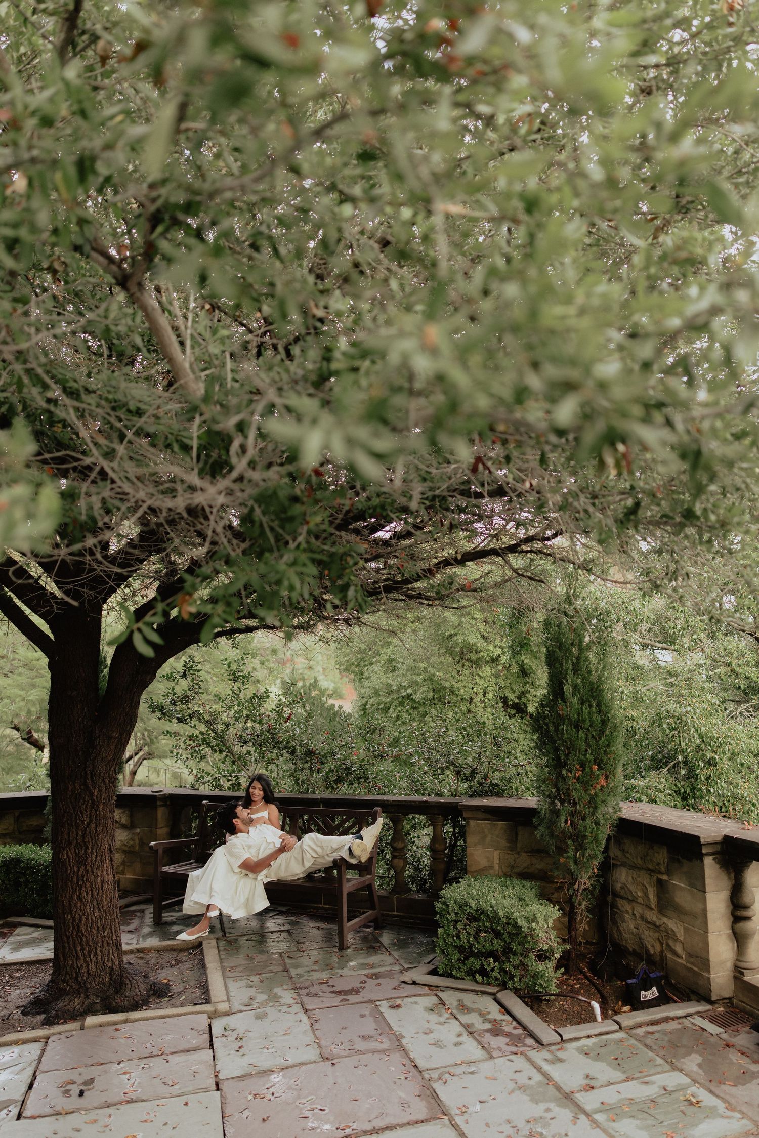 Bride in white gown sitting peacefully on a bench under a large tree in a lush garden patio.