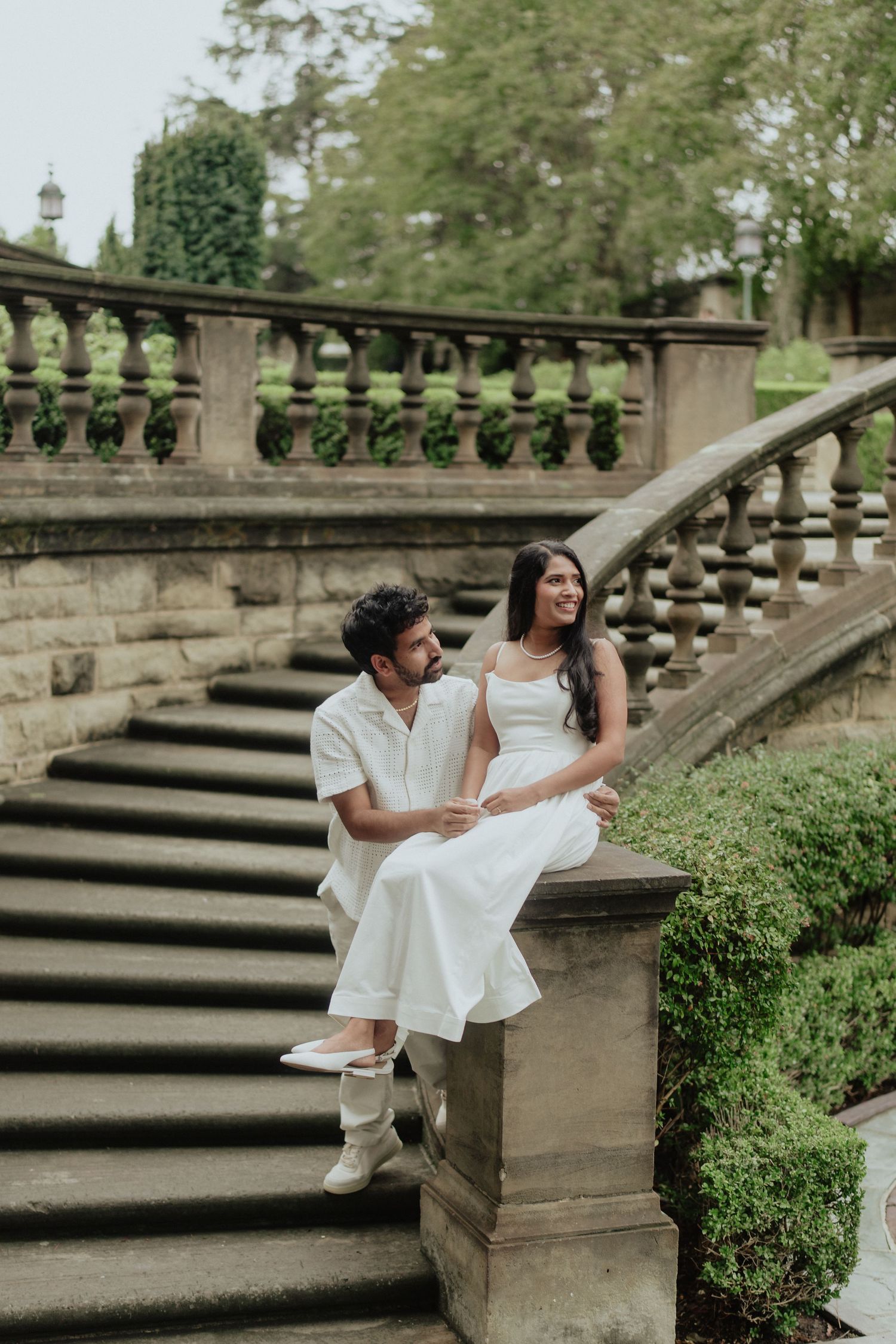Couple in white outfits sitting on stone balustrade steps in a lush garden setting.