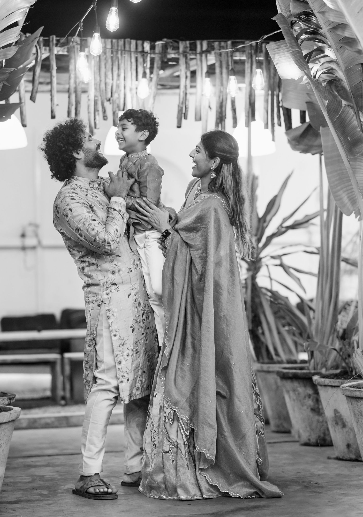 Black and white photo of a man in kurta holding a child while a woman in saree looks on, at a decorated venue.