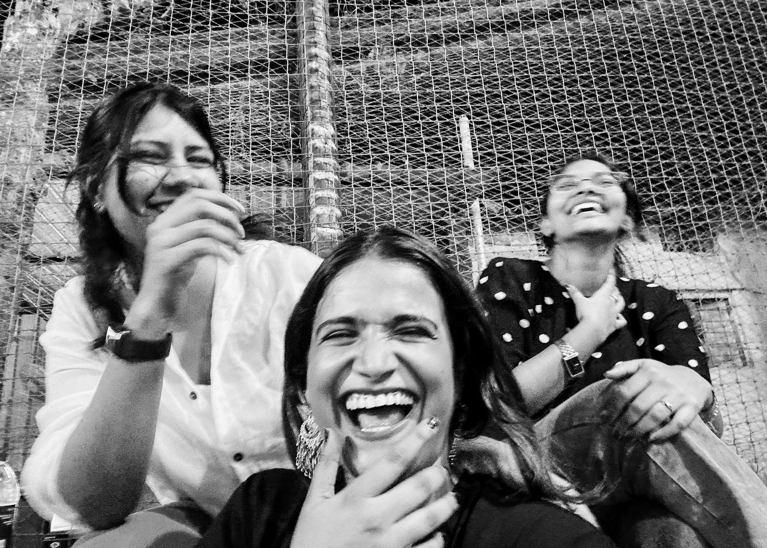 Three women laughing joyfully together in a black and white photo outdoors near a chain-link fence.