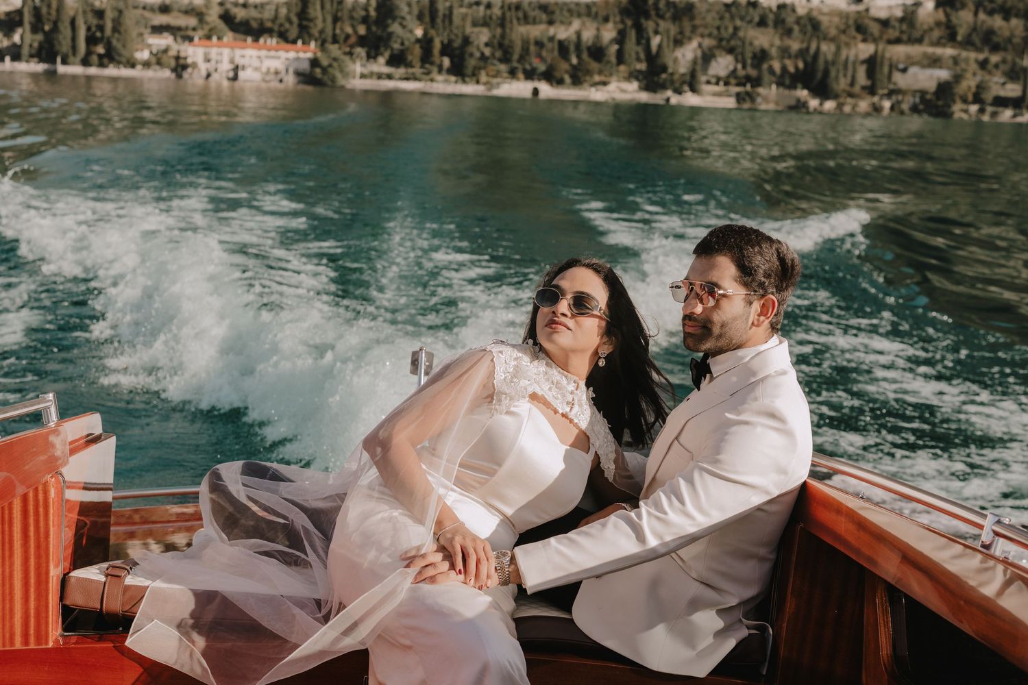 Bride and groom laughing on a wooden boat on turquoise water, bride in white dress and sunglasses.
