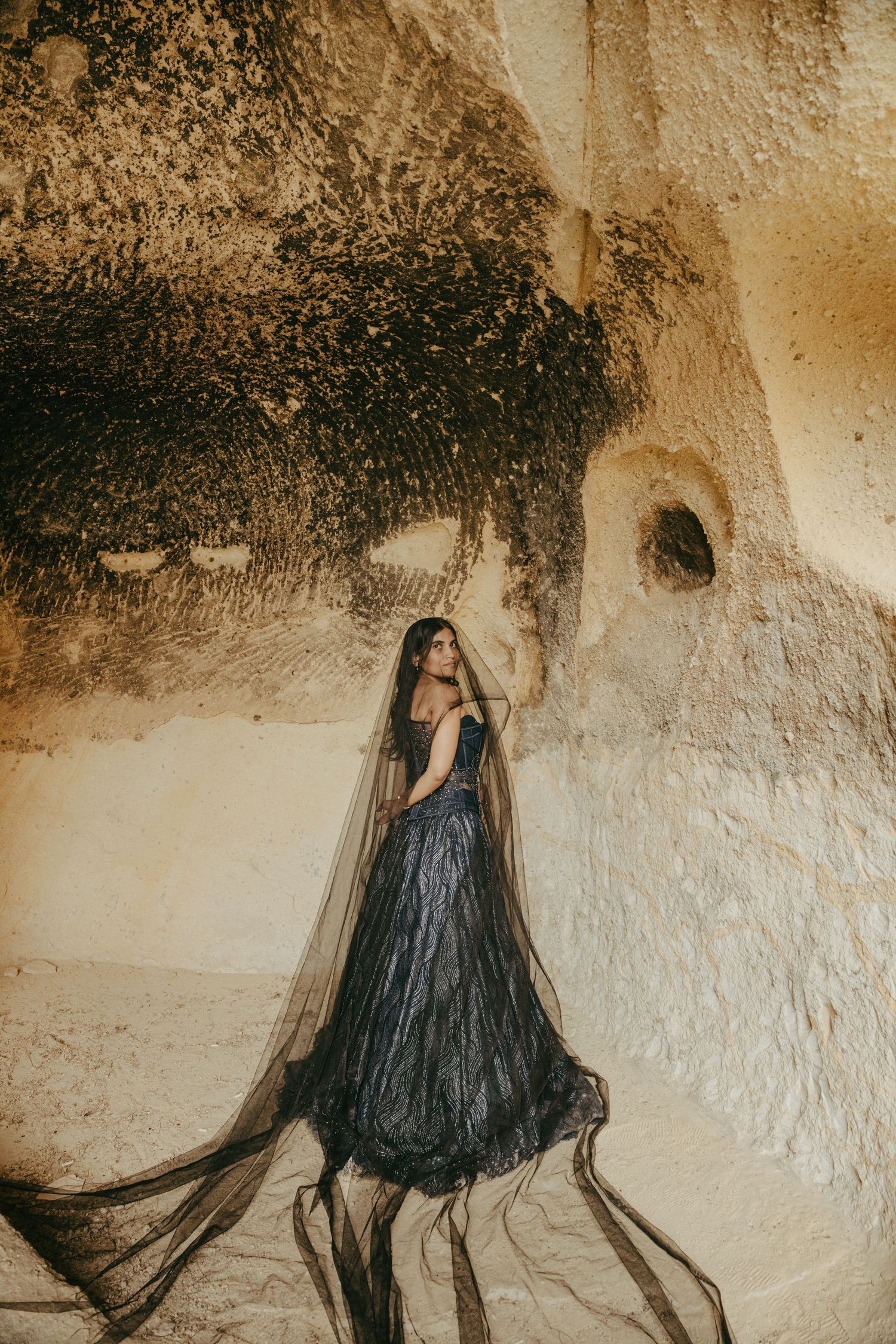 Bride in black gown with veil stands before ancient cave rock walls.