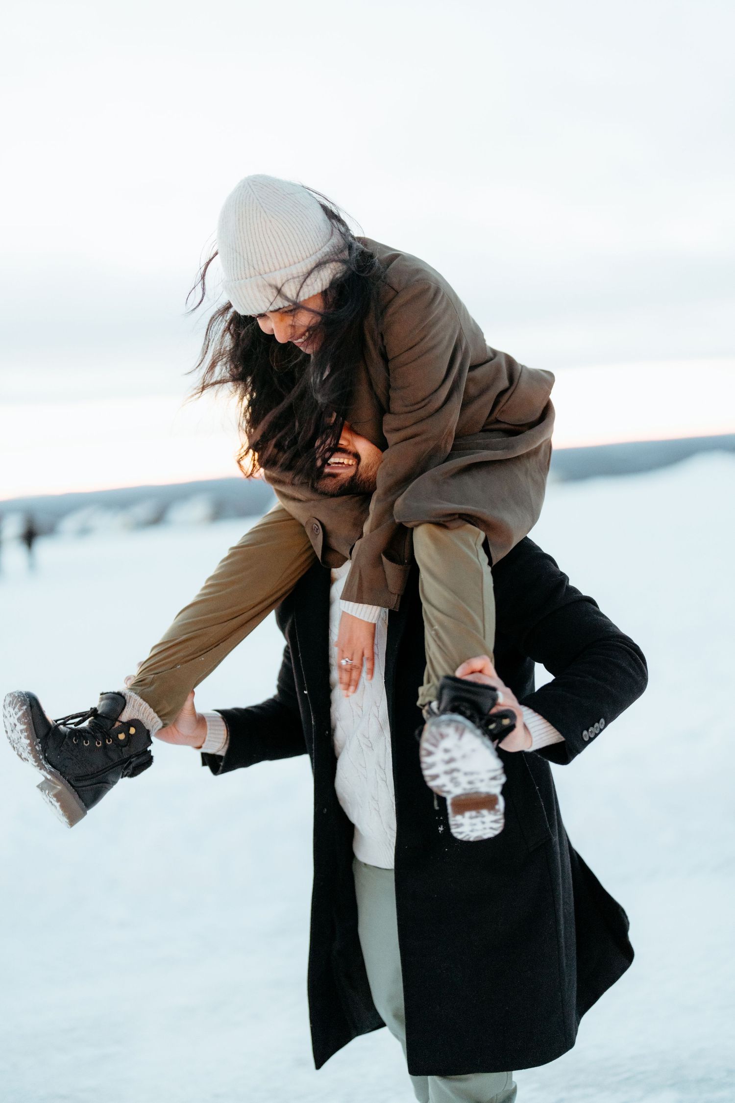 Couple playing in snow, man lifting woman on his shoulders, both wearing winter coats and boots outdoors.
