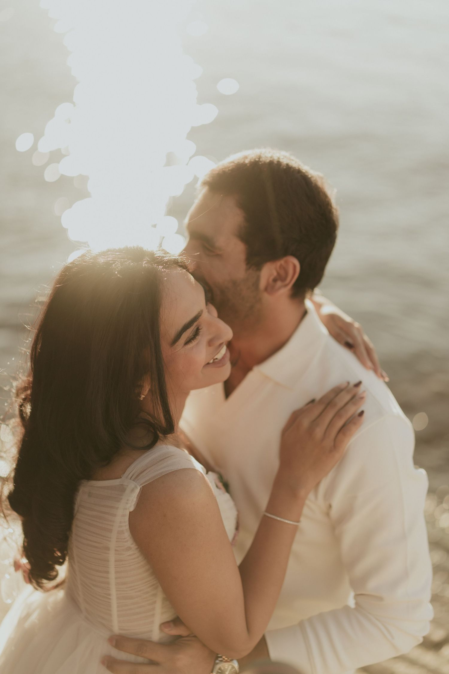 Romantic couple embracing at golden hour near water, woman smiling in white dress, man kissing her cheek in warm sunlight.