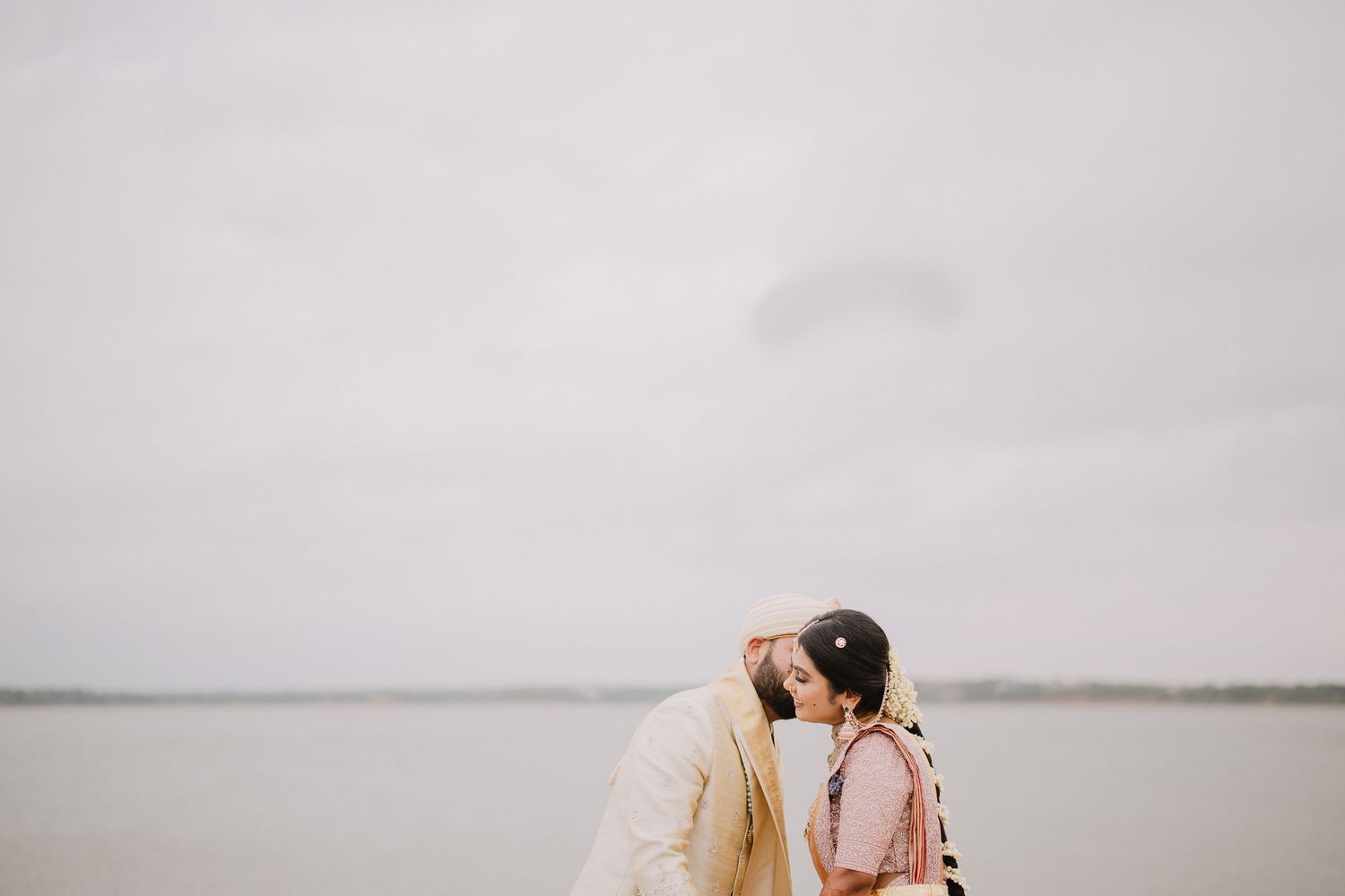 Couple sharing a romantic kiss by a serene waterfront on a cloudy day, dressed in elegant wedding attire.