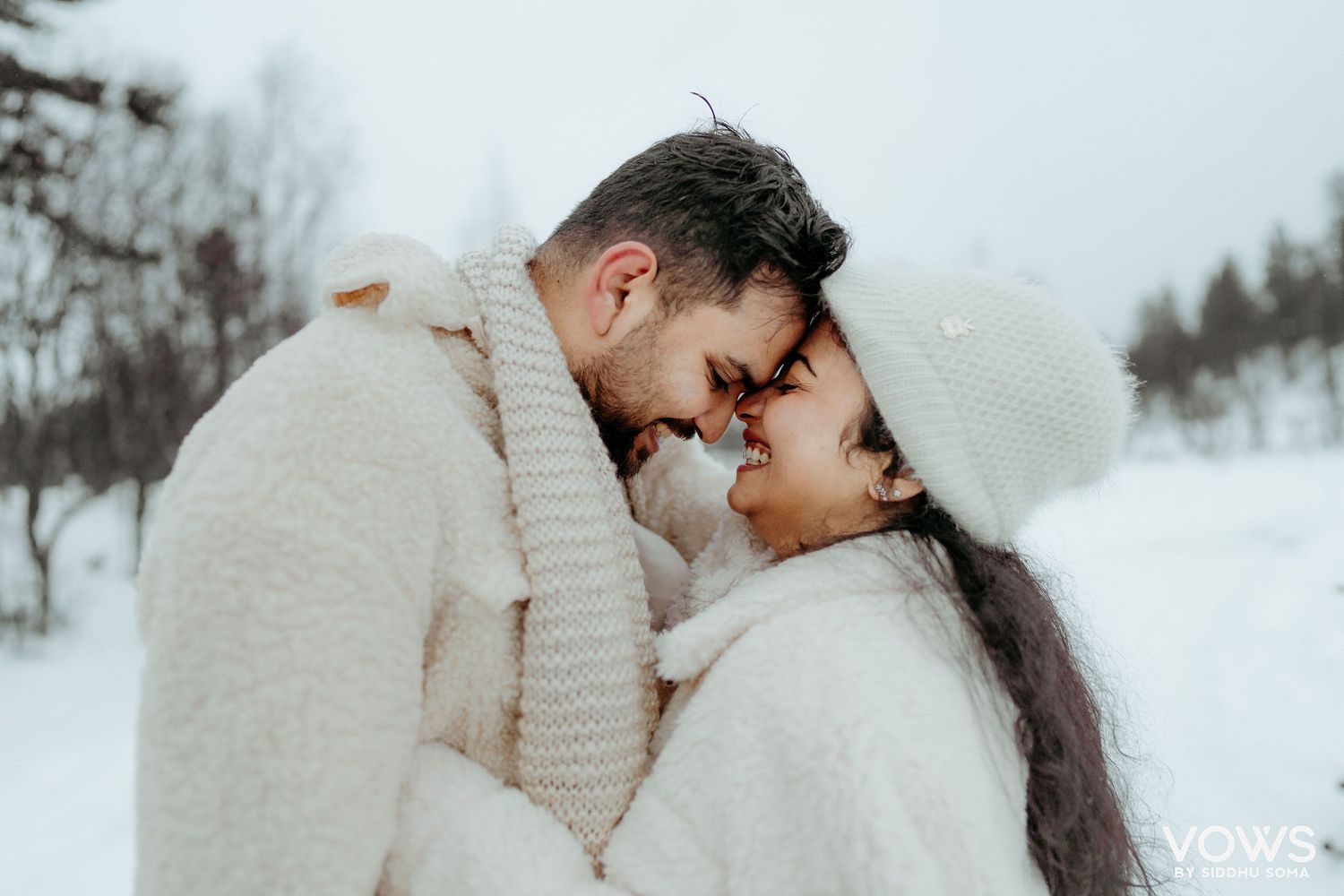 Romantic couple in matching white winter sweaters touching foreheads in snowy landscape.