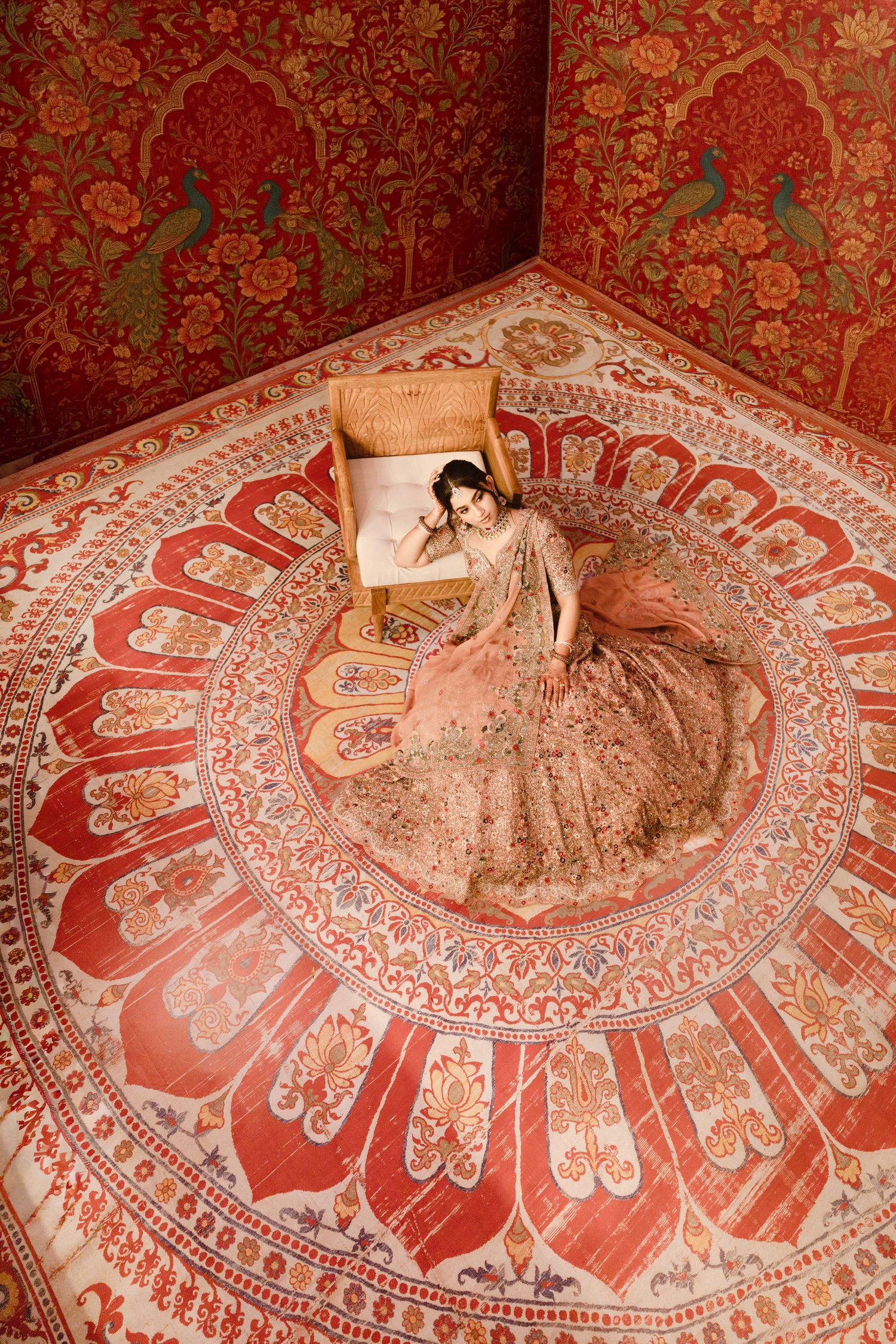 Woman in elegant peach lehenga seated on ornate carpet with red floral walls in traditional Indian setting.