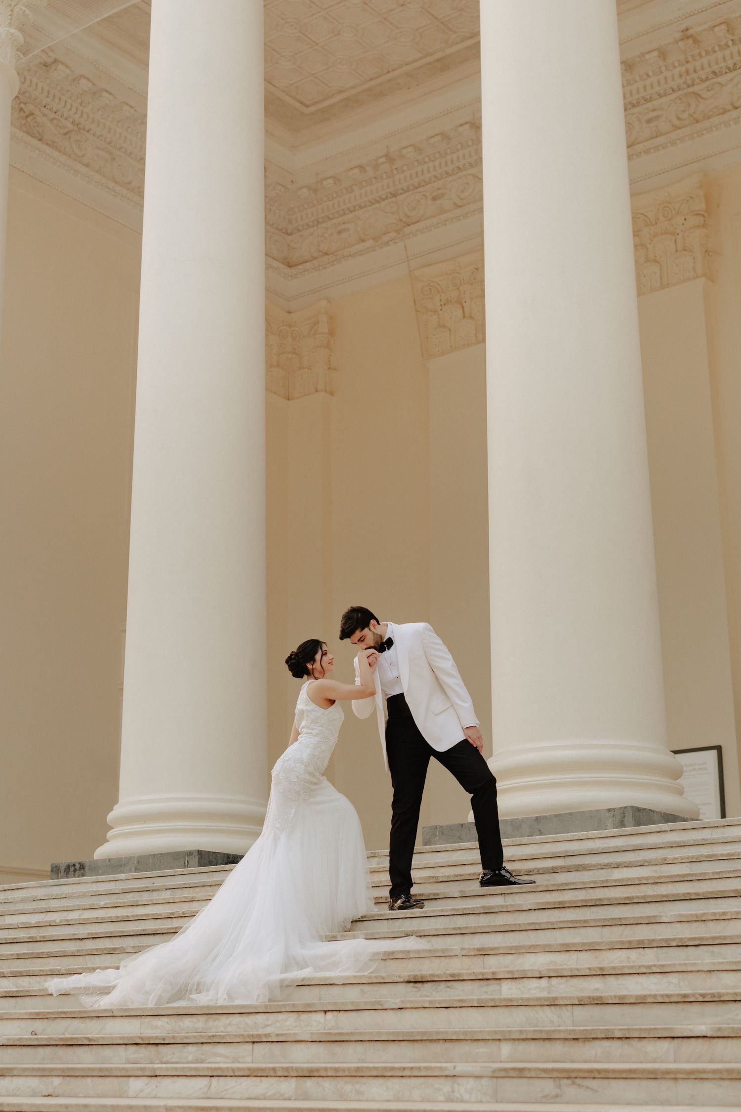 Bride and groom share a romantic moment on marble steps between towering white columns.