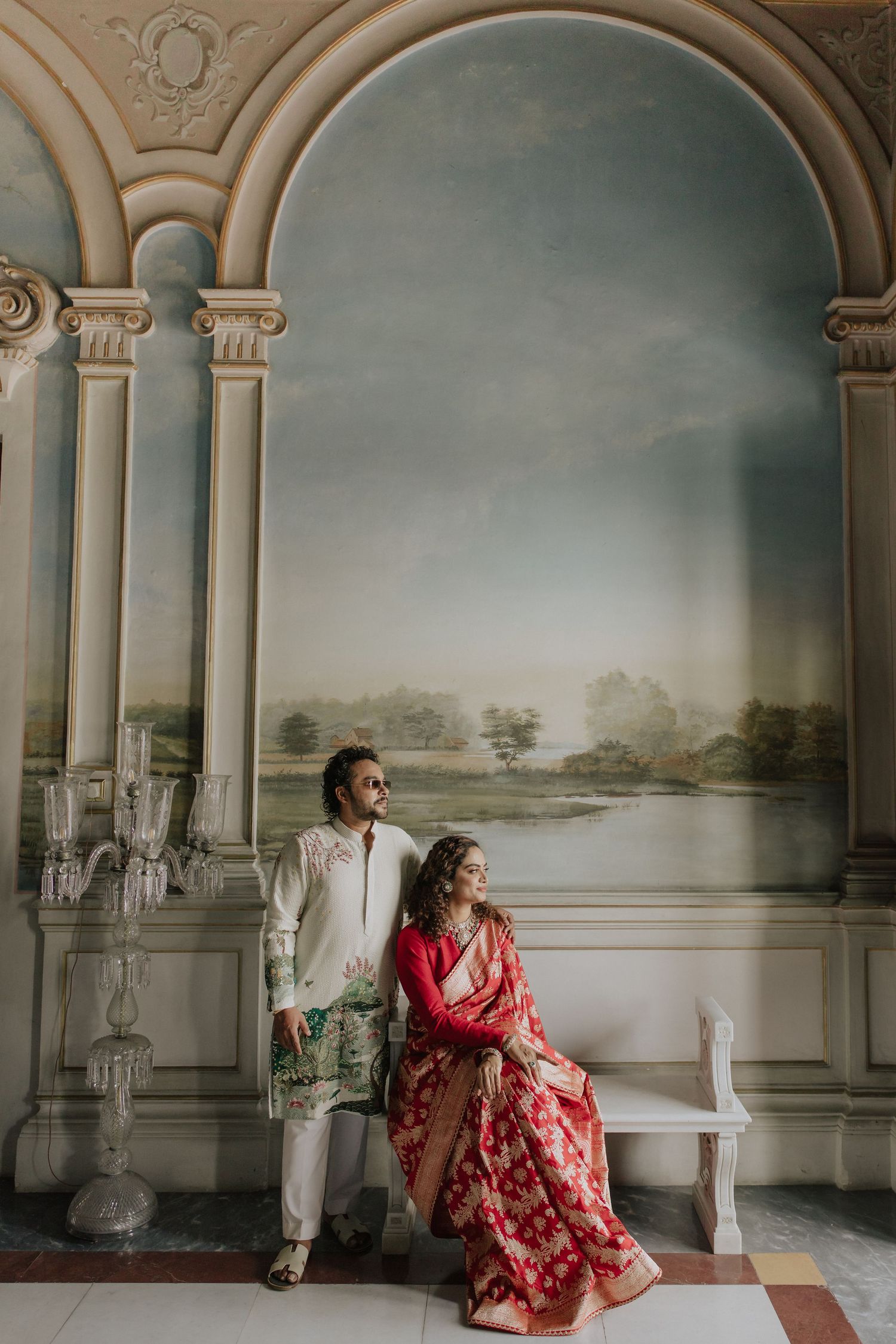Indian couple in traditional wedding attire posing before an ornate mural with classical architectural details.