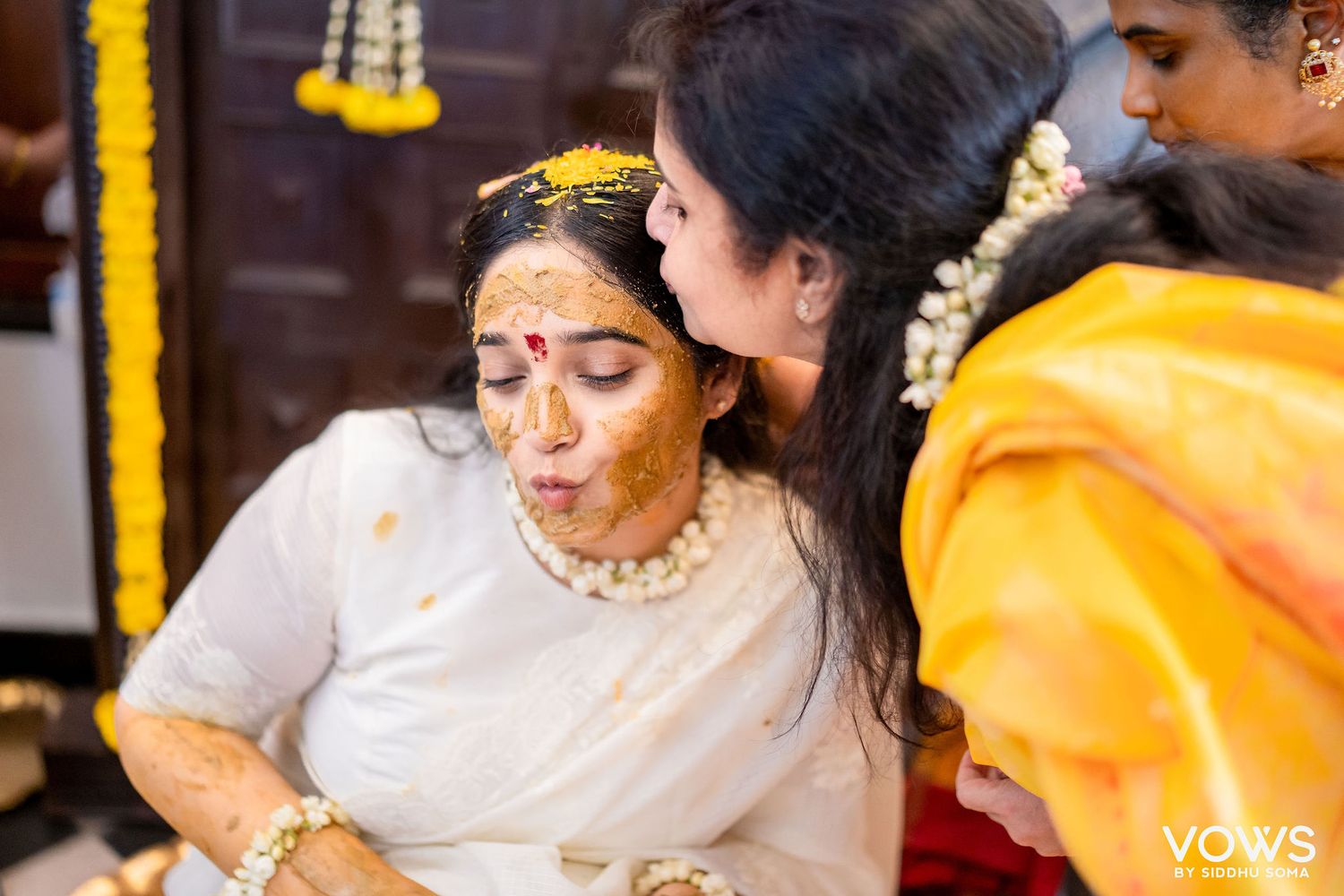 A bride in white saree gets turmeric haldi applied on her face during a traditional South Indian wedding ceremony.