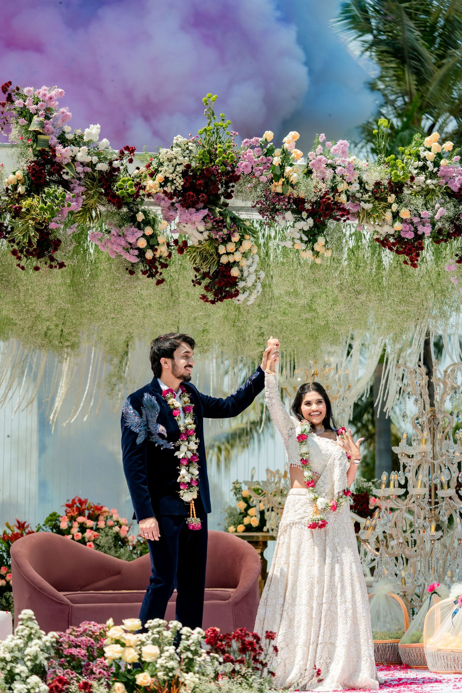 Indian couple celebrating under floral arch with colorful smoke at luxurious wedding ceremony venue.