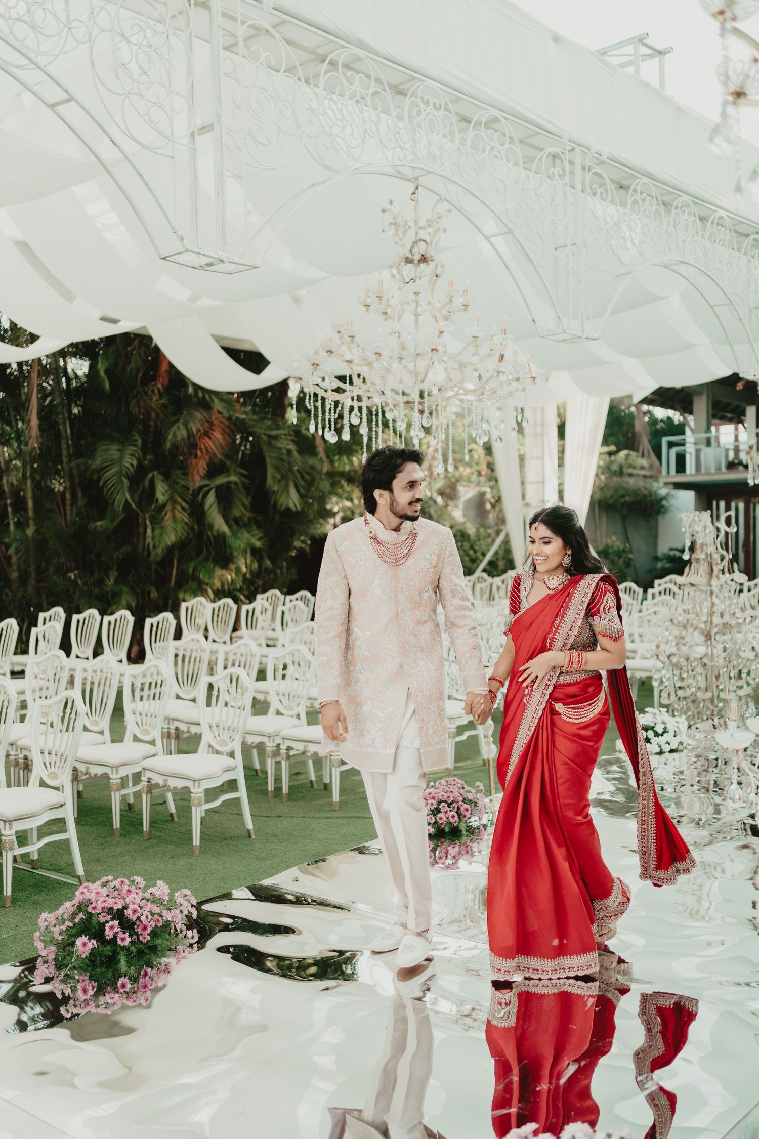 Indian bride in red saree and groom in cream sherwani walking down aisle at elegant wedding venue.